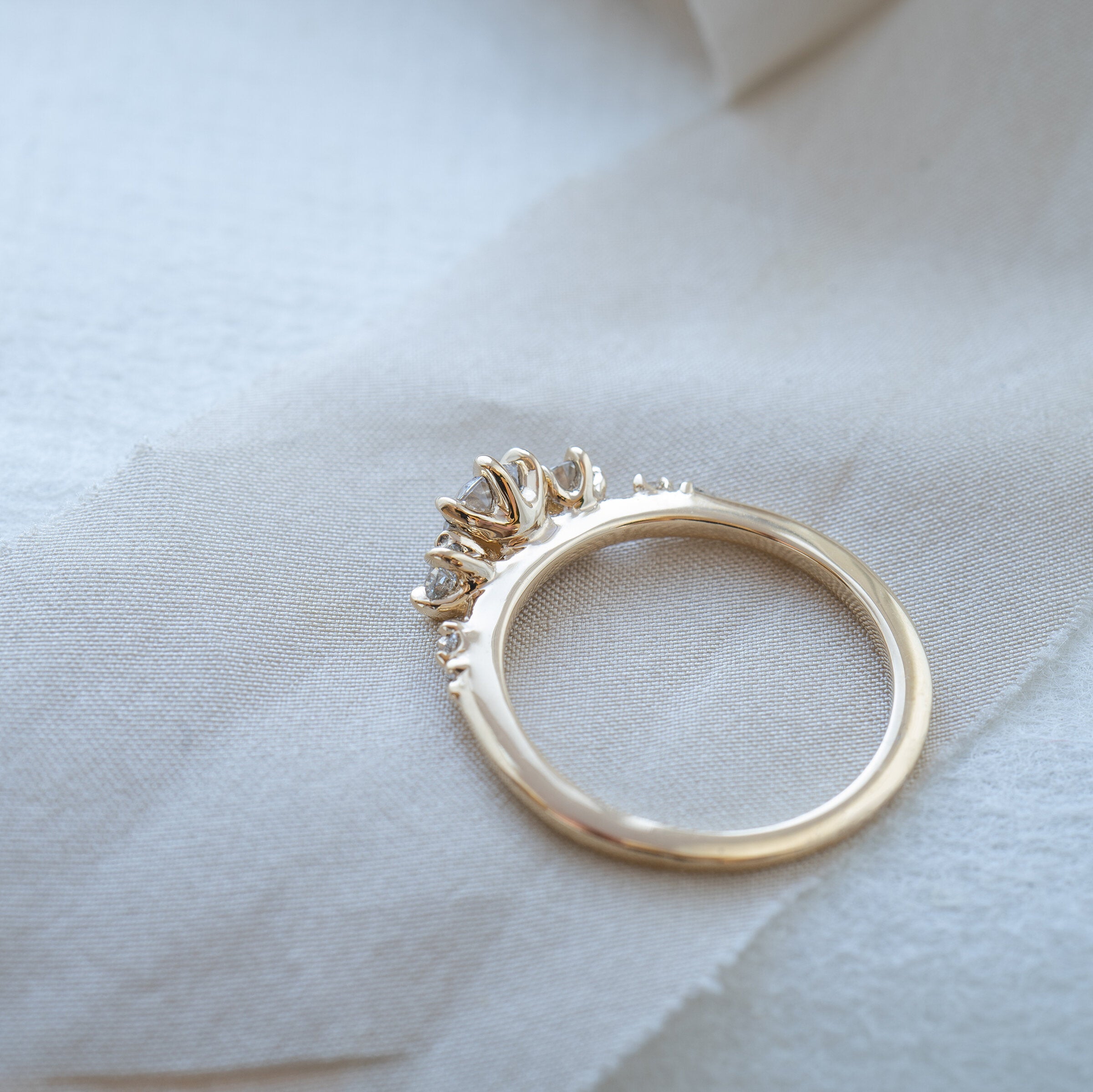 A Wild Iris three stone Engagement Ring by Laurie Fleming Jewellery, lying flat on a piece of raw off-white ribbon. The angle shows the side view of the ring, including its smooth solid gold band and its flat, plain sides which let it stack beautifully with other rings.