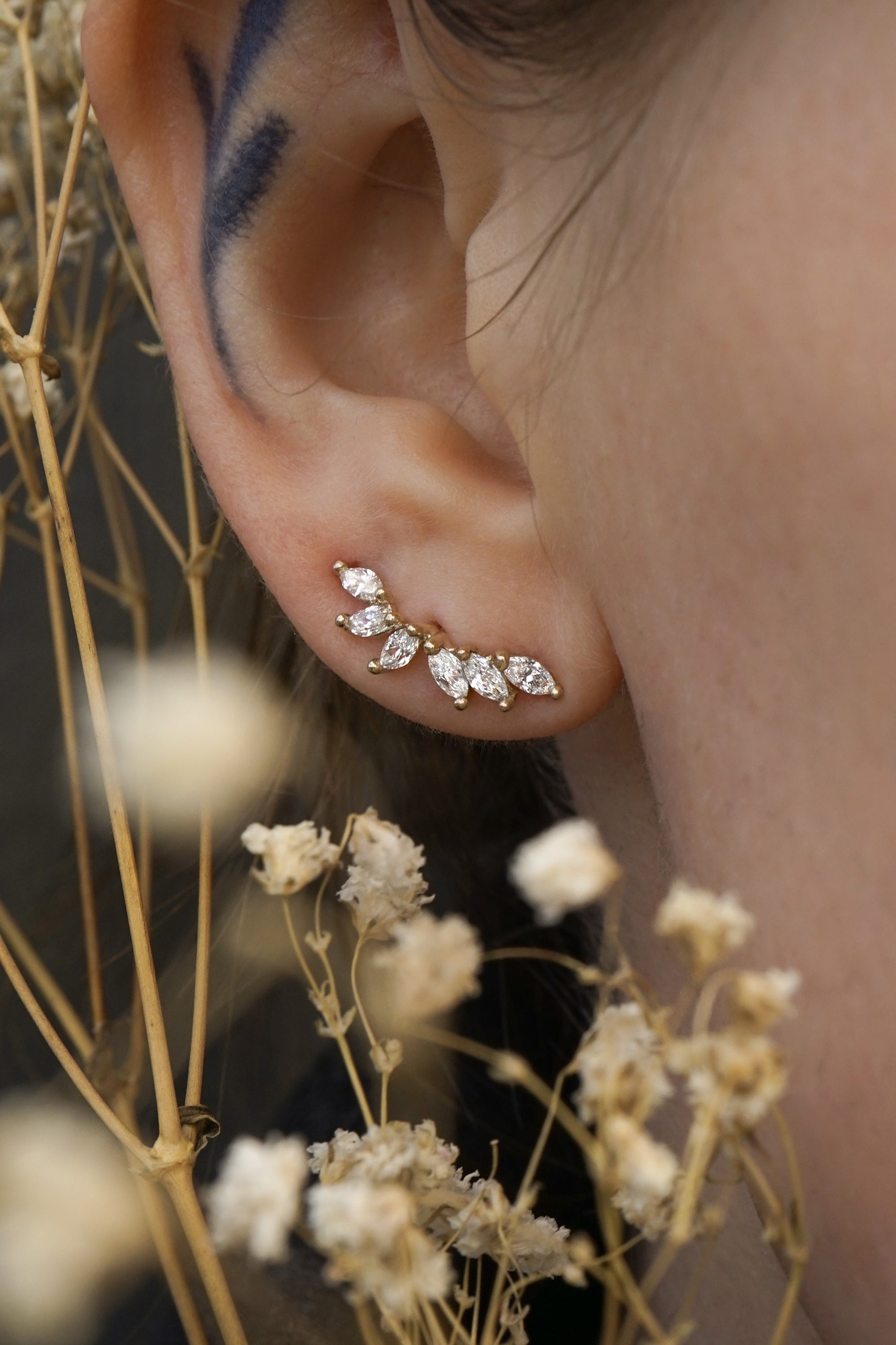 A close up of an ear wearing a Lyrie stud earring with six marquise cut diamonds. There are dried flower stems in the foreground around the ear.