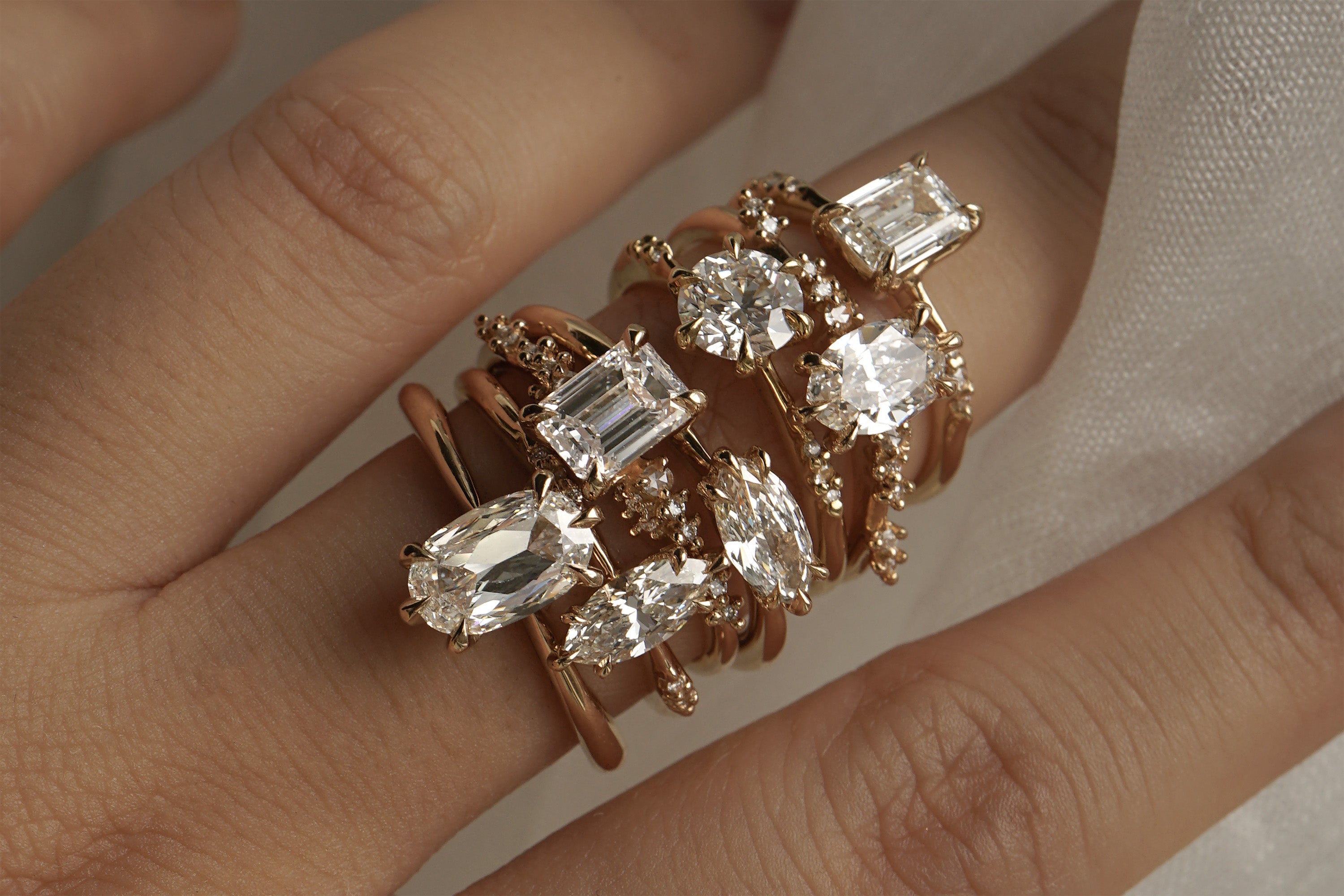 A hand with a stack of diamond engagement rings by Laurie Fleming Jewellery on the middle finger. From top to bottom: Ilona ring with emerald cut diamond, Daphne Ring with oval cut diamond, Ilona ring with round brilliant cut diamond, Aurelia ring with a moval (marquise/oval) diamond, Daphne ring with an emerald cut diamond, Ilona ring with a marquise cut diamond, Aurelia ring with an oval cut diamond.