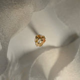 A product photo of a Pansy stud by Laurie Fleming Jewellery. It features a hand-carved miniature Pansy flower in solid 14k gold with a round brilliant cut diamond centre. The diamond can be swapped out for any birthstone. The background is a light grey with bits of raw silk at the top and bottom of the frame.