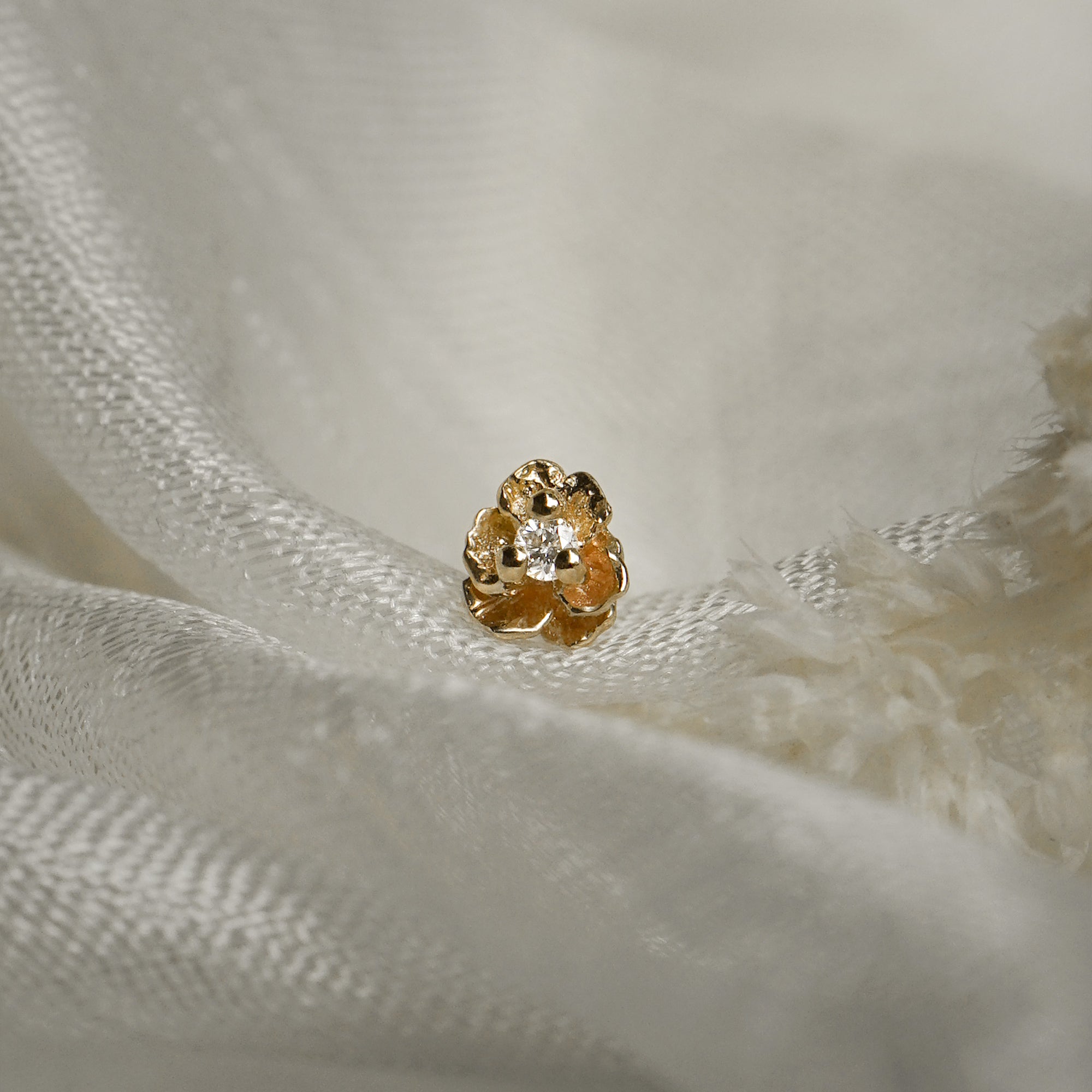 A product photo of a Pansy stud by Laurie Fleming Jewellery. It features a hand-carved miniature Pansy flower in solid 14k gold with a round brilliant cut diamond centre. The diamond can be swapped out for any birthstone. The stud is resting on some raw white silk.