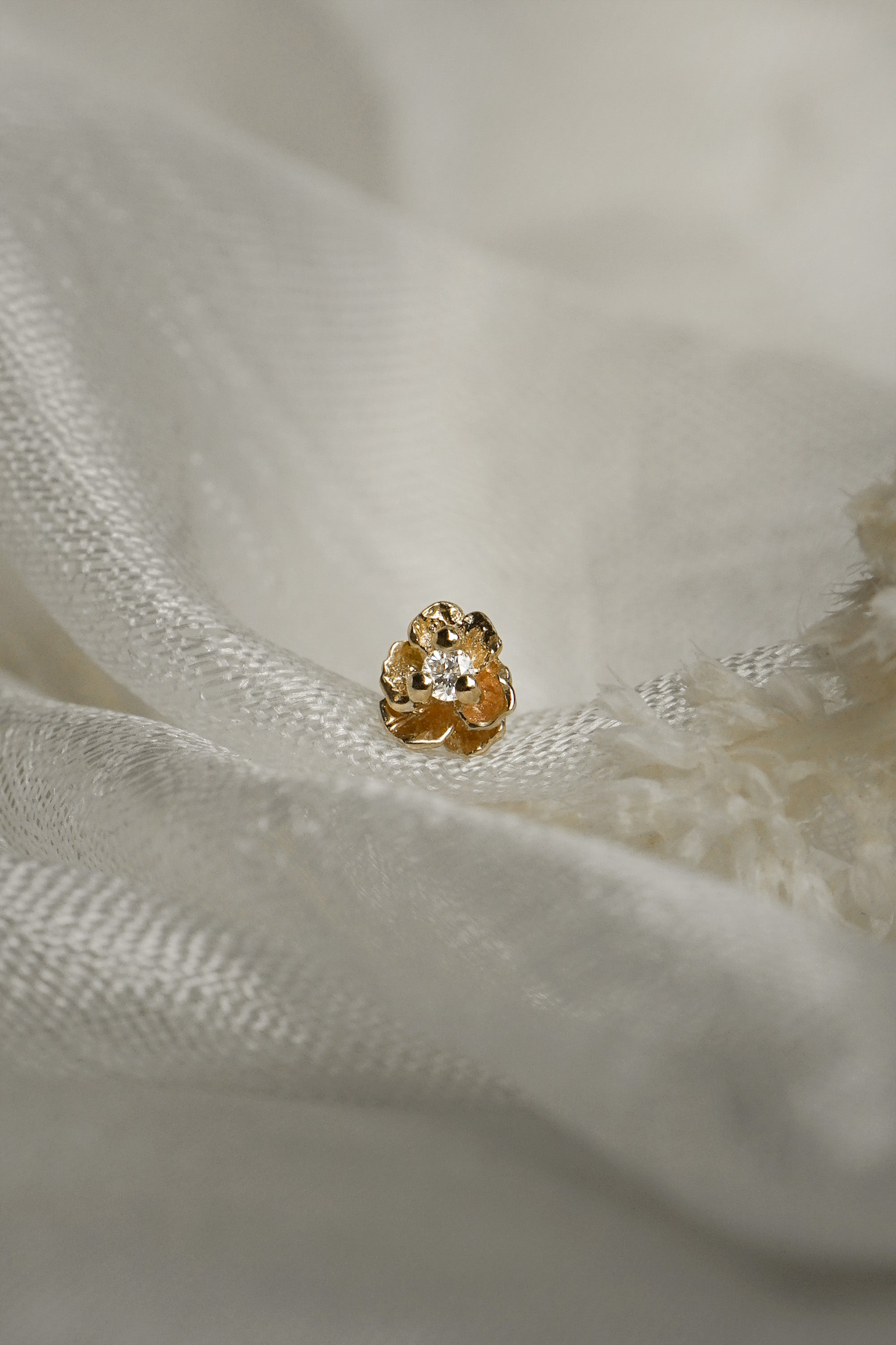A product photo of a Pansy stud by Laurie Fleming Jewellery. It features a hand-carved miniature Pansy flower in solid 14k gold with a round brilliant cut diamond centre. The diamond can be swapped out for any birthstone. The stud is resting on some raw white silk.