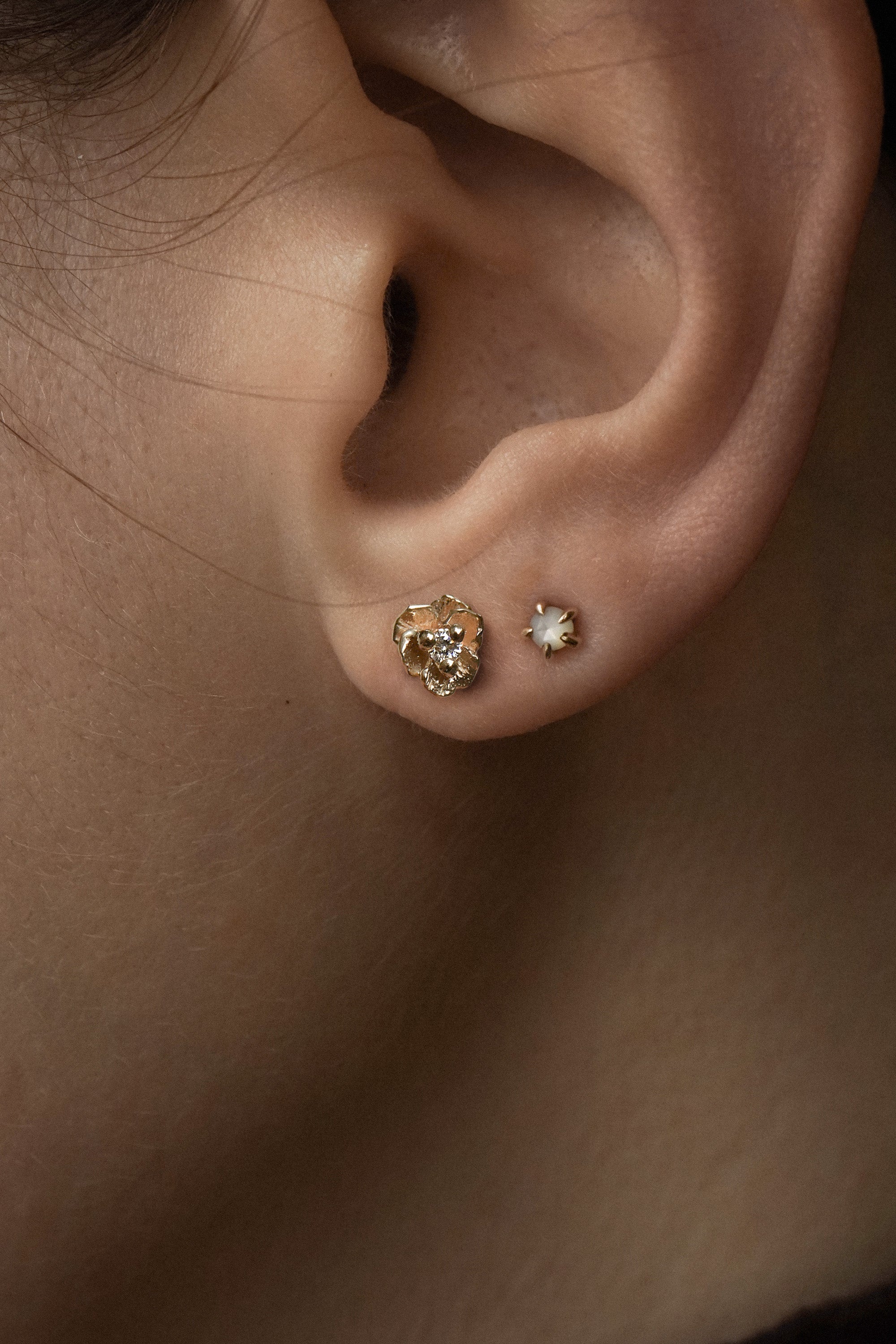 A close-up of an ear wearing two stud earrings by Laurie Fleming Jewellery. The model has tan skin and both earrings are in lobe piercings. The bottom piercing features a diamond Pansy stud, and the top piercing features a rose cut mother of pearl moondew stud.