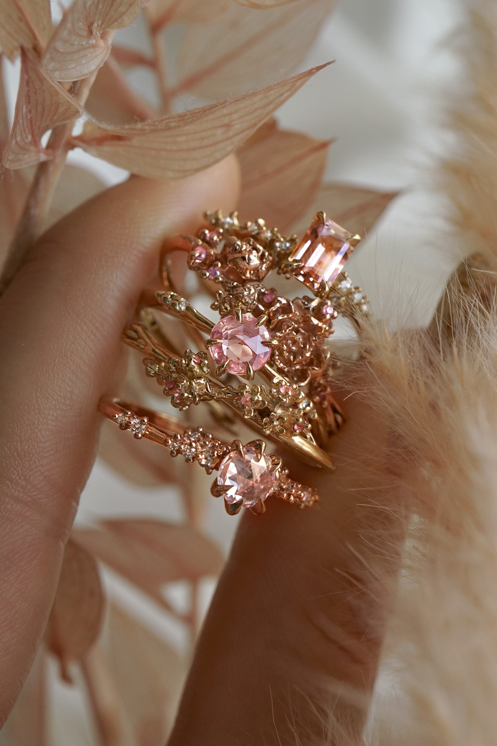 A photo of three rings and two hoops made by Laurie Fleming Jewellery held between index finger and thumb, including:  A "Daphne" style engagement ring featuring an emerald cut peach sapphire, two one of a kind "Asrai Garden" hoops featuring hand-carved miniature flowers, an "Ilona" style engagement ring with a round rose cut pink sapphire, and a "Daphne" style engagement ring with a round rose cut pale pink sapphire. There are leaves and feathers in shades of soft beige and pink surrounding the hand,