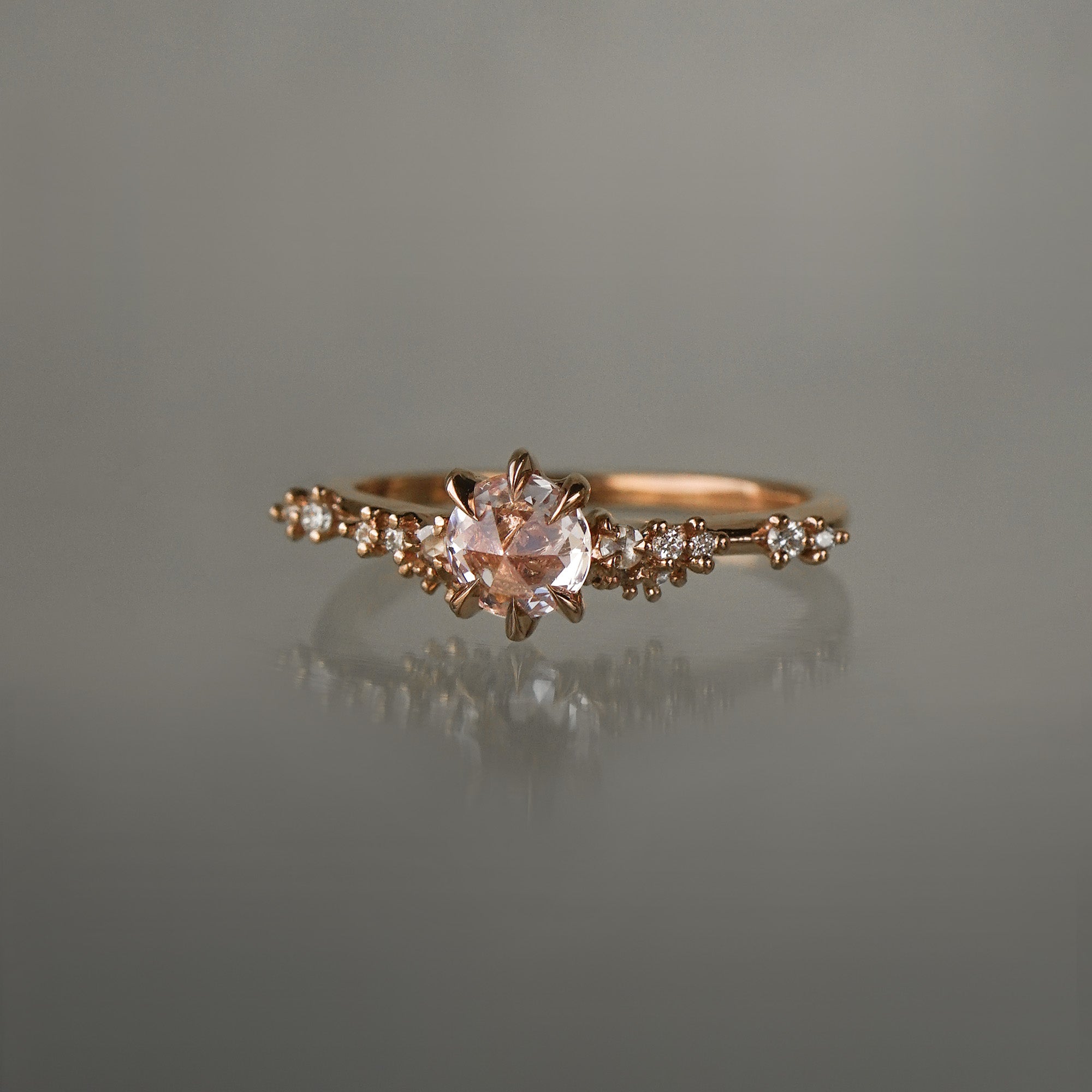 A delicate and ethereal hand-carved one of a kind "Daphne" style engagement ring by Laurie Fleming Jewellery. The ring features a rose cut pale pink sapphire centre, with clusters of rose and brilliant cut diamond accents on the band in solid 14k rose gold. The ring is situated against a medium grey background.