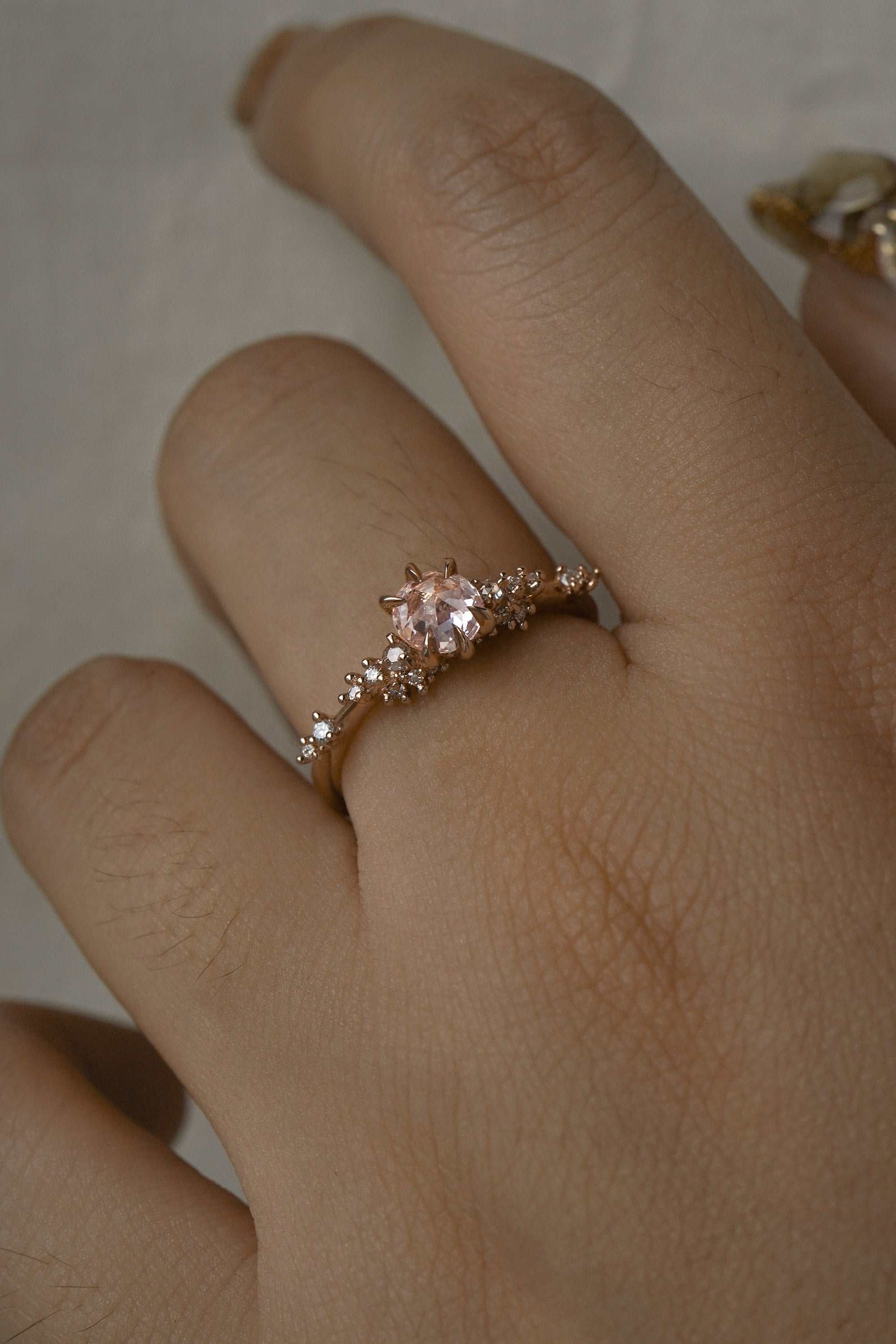 A delicate and ethereal hand-carved one of a kind "Daphne" style engagement ring by Laurie Fleming Jewellery. The ring features a rose cut pale pink sapphire centre, with clusters of rose and brilliant cut diamond accents on the band in solid 14k rose gold. The ring is worn on a hand against a light grey background.