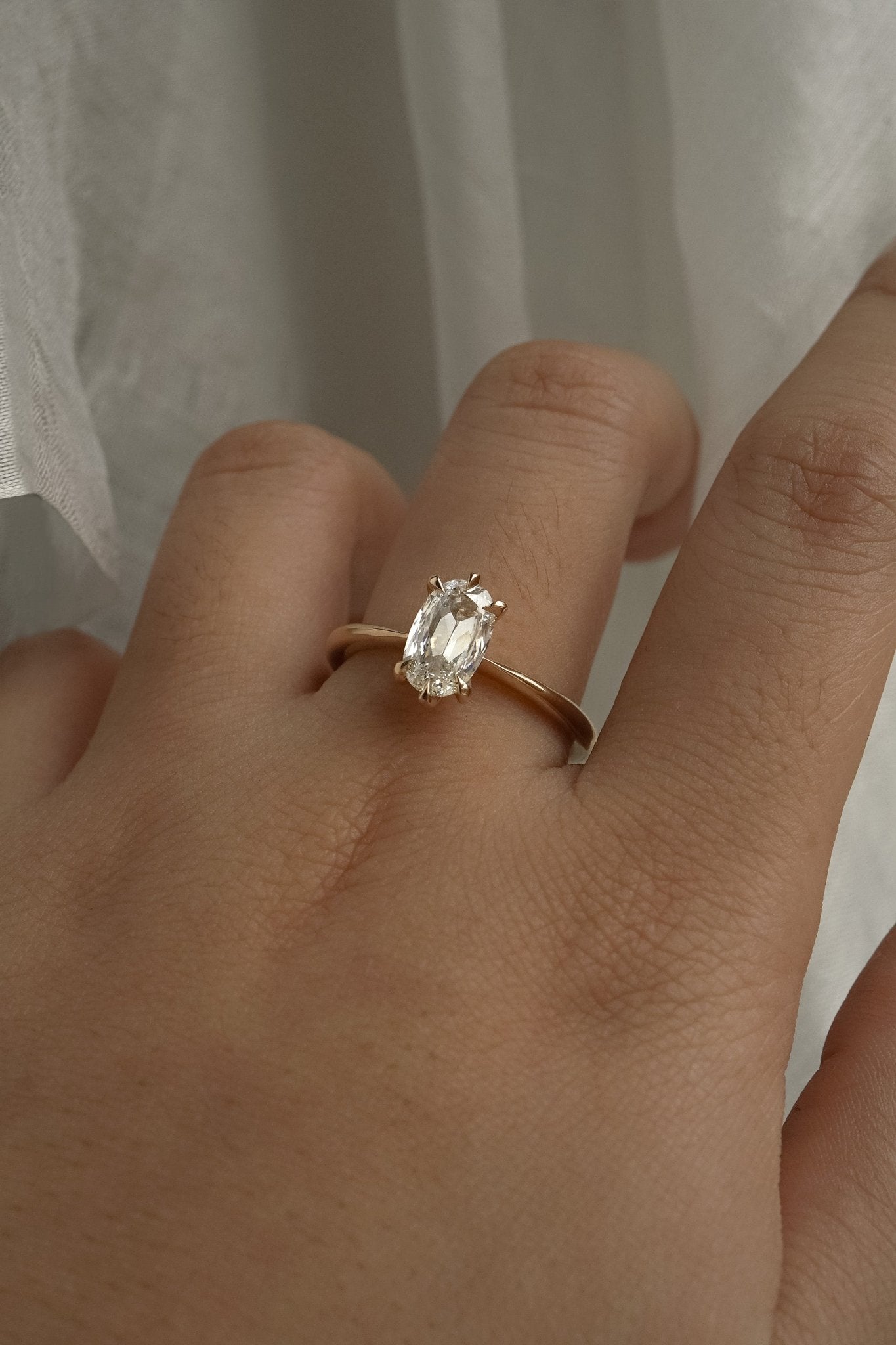 A one of a kind "Aurelia" style ring by Laurie Fleming Jewellery, featuring a unique elongated oval diamond with antique-inspired facets, on a gently tapered and rounded solid gold band. The ring is worn on a hand posed in front of some draped gossamer silk fabric.