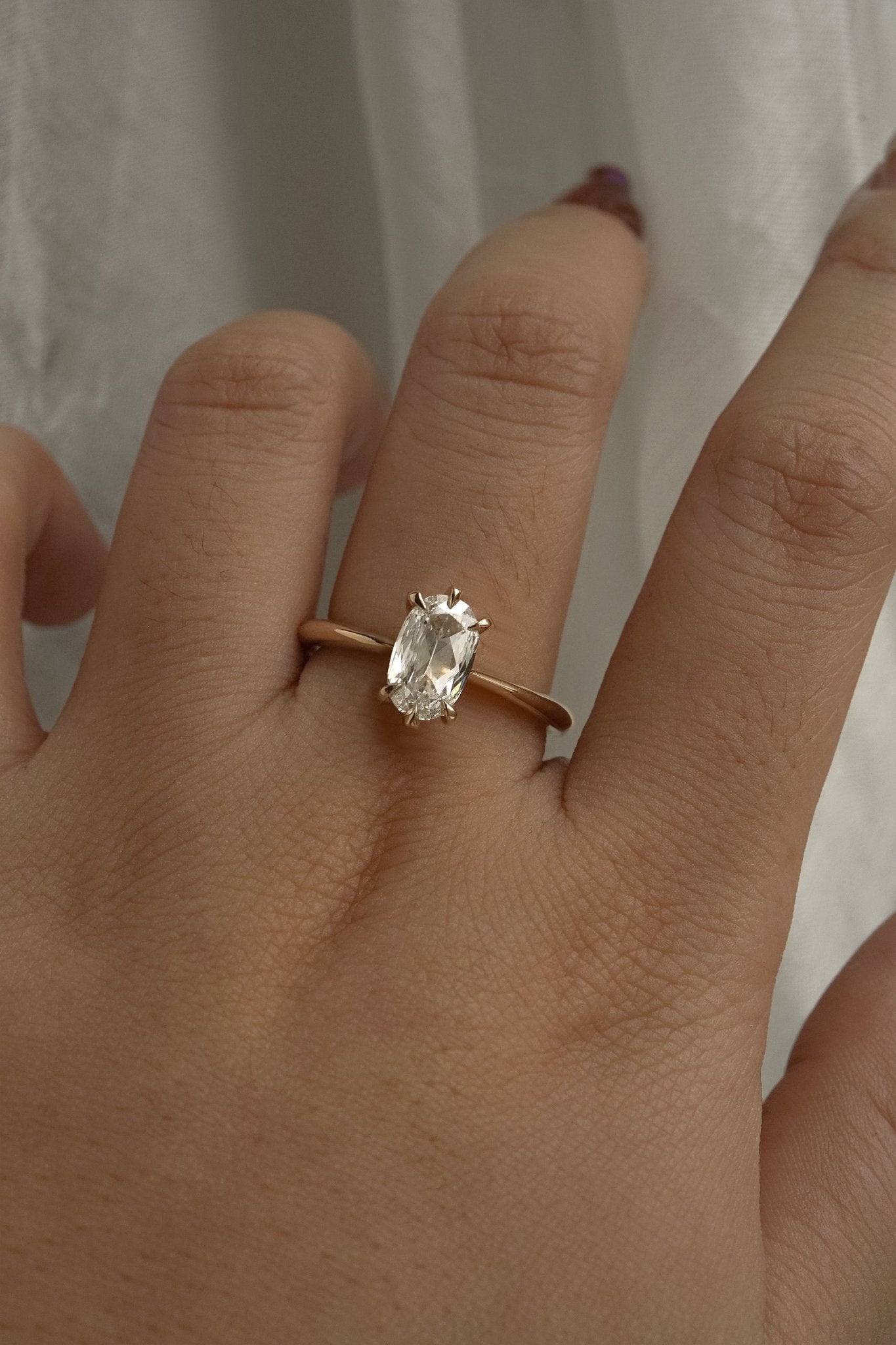 A one of a kind "Aurelia" style ring by Laurie Fleming Jewellery, featuring a unique elongated oval diamond with antique-inspired facets, on a gently tapered and rounded solid gold band. The ring is worn on a hand posed in front of some draped gossamer silk fabric.