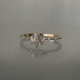 A one of a kind 14k champagne gold "Water Lily" engagement ring by Laurie Fleming Jewellery with a silver-blush colour change Montana sapphire centre held in delicate hand-carved claw prongs. A sprinkling of petite and delicate diamonds dance along the band. The ring is situated on a medium grey background.
