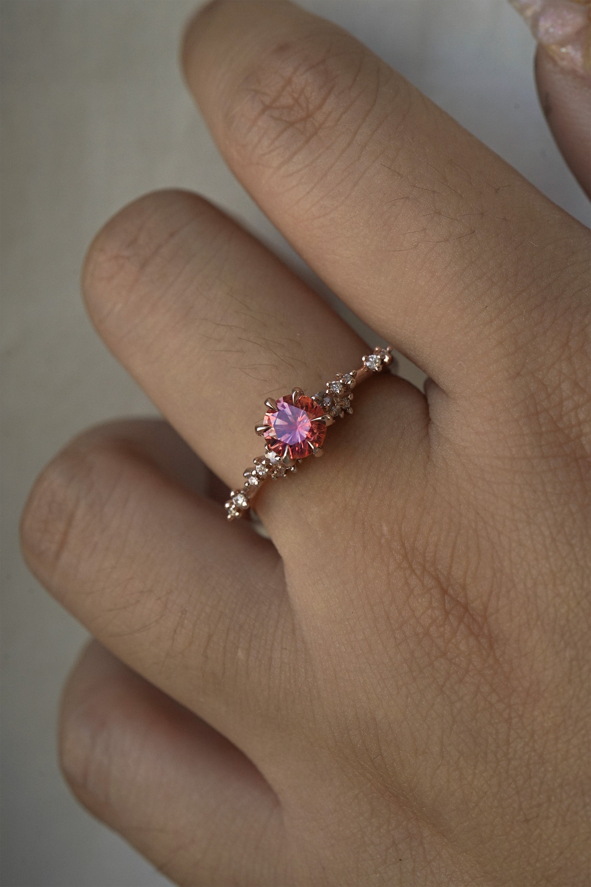 A delicate and ethereal hand-carved one of a kind "Daphne" style engagement ring by Laurie Fleming Jewellery. The ring features a round brilliant cut red peach padparascha sapphire centre, with clusters of rose and brilliant cut diamond accents on the band in 14k rose gold. The ring is worn on a hand against a light grey background.