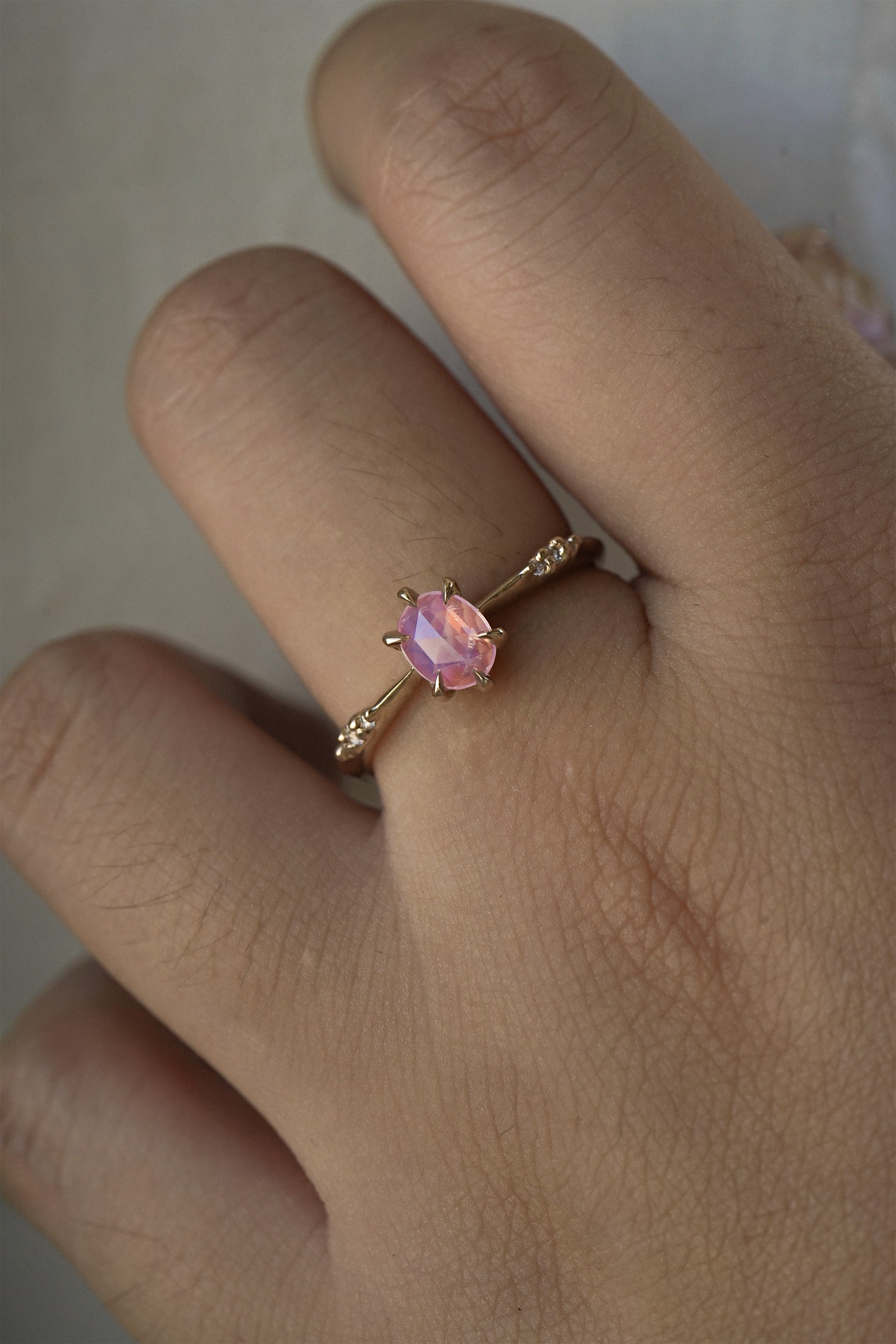 A delicate hand-carved one of a kind "Ilona" style engagement ring by Laurie Fleming Jewellery. The ring features a silky and opalescent rose cut oval pink sapphire centre, a tapered knife-edge band, and small diamond accents on the band shoulders. The ring is worn on a hand against a light grey background.