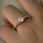 A one of a kind "Daphne" style engagement ring by Laurie Fleming Jewellery, worn on a hand in front of a light grey plain background.  The ring features a pale rosewater pink sapphire centre. The band is encrusted in sparkling diamond clusters. The ring is handcarved and made in solid champagne yellow gold.