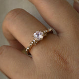 A one of a kind "Daphne" style engagement ring by Laurie Fleming Jewellery, worn on a hand in front of a light grey plain background.  The ring features a pale rosewater pink sapphire centre. The band is encrusted in sparkling diamond clusters. The ring is handcarved and made in solid champagne yellow gold.