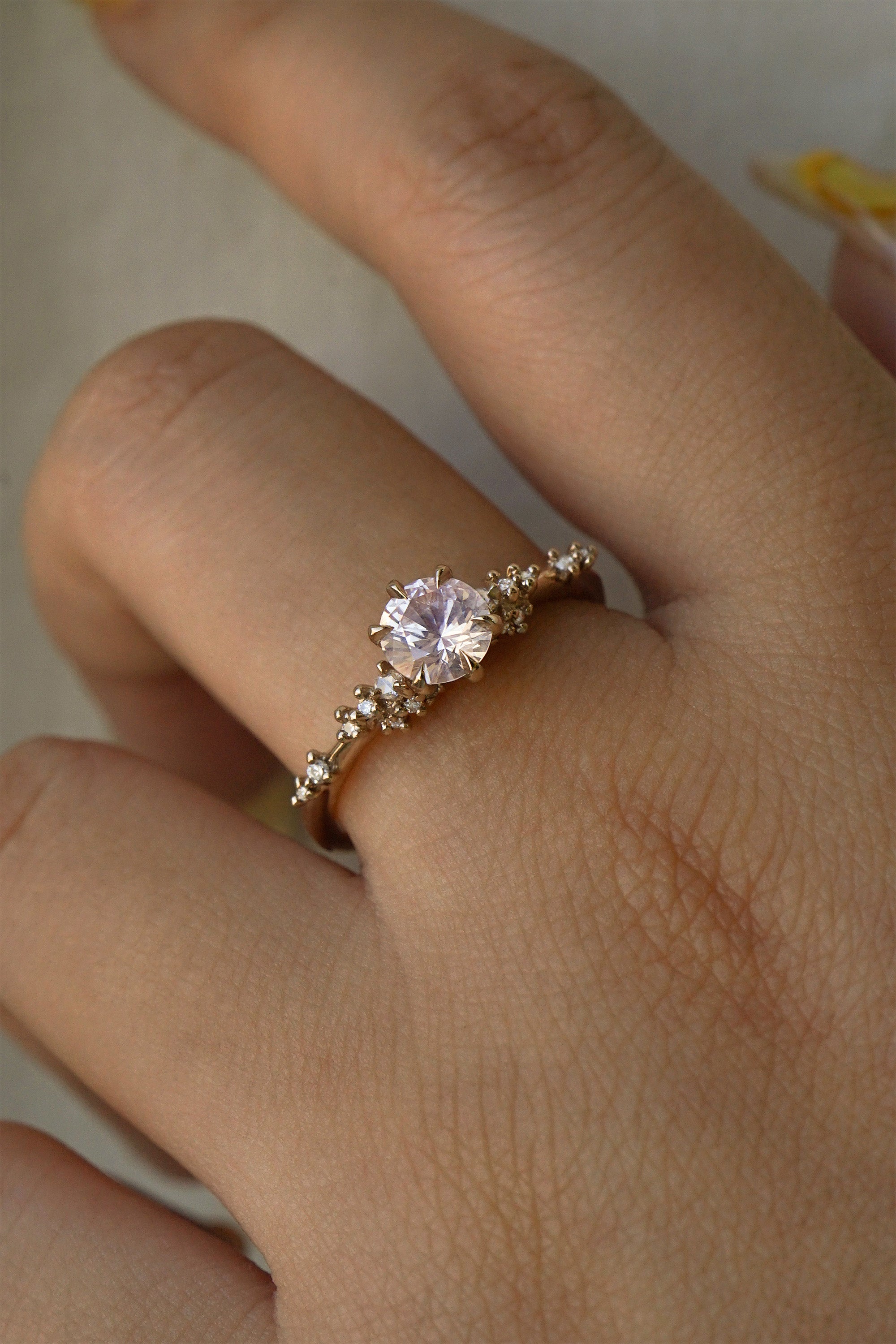 A one of a kind "Daphne" style engagement ring by Laurie Fleming Jewellery, worn on a hand in front of a light grey plain background.  The ring features a pale rosewater pink sapphire centre. The band is encrusted in sparkling diamond clusters. The ring is handcarved and made in solid champagne yellow gold.