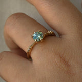 Dainty yellow gold Ilona style one of a kind engagement ring by Laurie Fleming Jewellery, worn on a hand in front of a light grey background. The ring features a silky seafoam turquoise green sapphire and delicate twinkling diamonds on the band. 