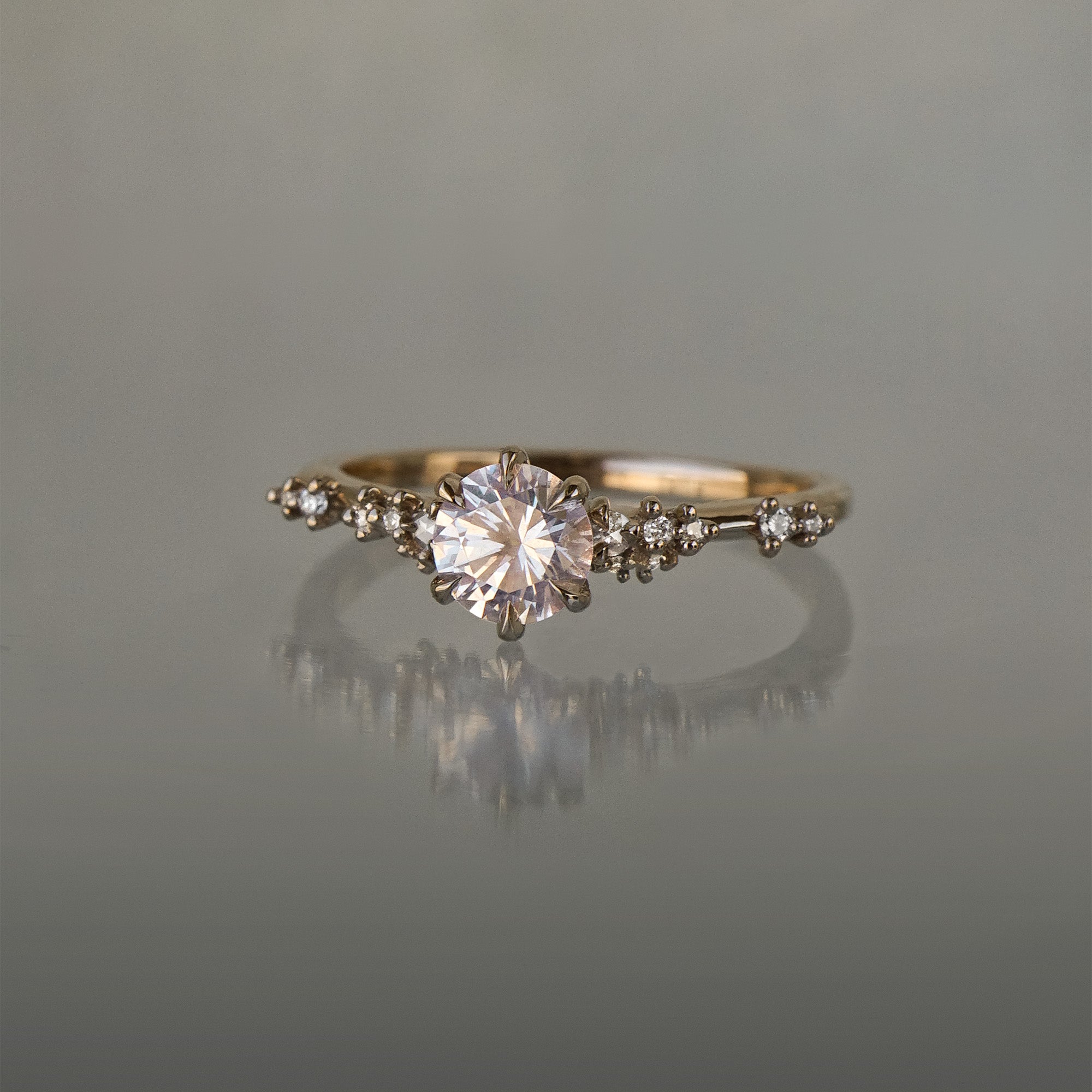 A one of a kind "Daphne" style engagement ring by Laurie Fleming Jewellery featuring a pale rosewater pink sapphire centre. The band is encrusted in sparkling diamond clusters. The ring is handcarved and made in solid champagne yellow gold, and is situated on a medium grey background.