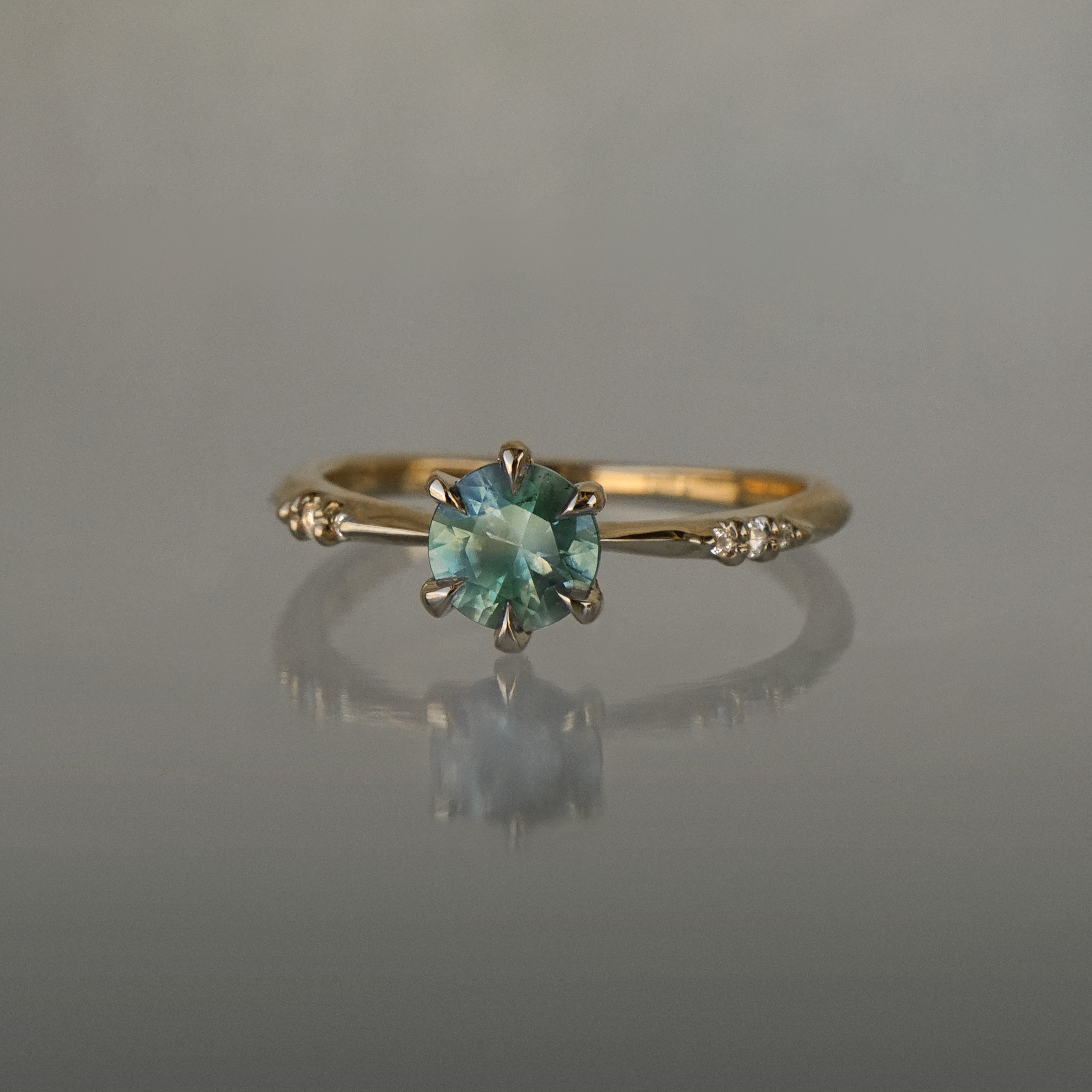 Dainty yellow gold Ilona style one of a kind engagement ring by Laurie Fleming Jewellery on a grey background, with a silky seafoam turquoise green sapphire and delicate twinkling diamonds on the band.