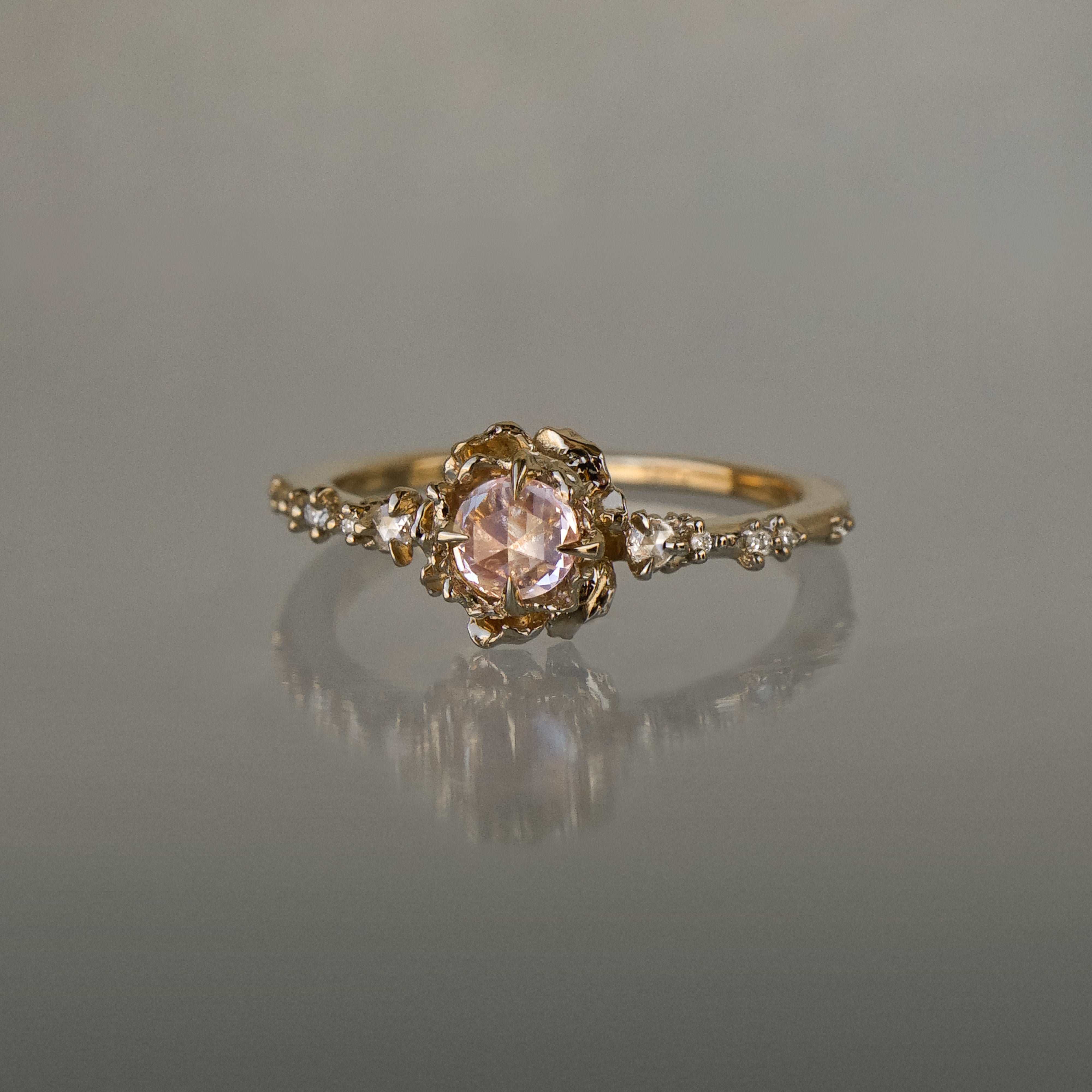 A delicate, one of a kind "Nereid" style engagement ring by Laurie Fleming Jewellery in yellow gold. The ring features a round rose cut light pink sapphire held by four dainty claws, nestled in hand-carved floral peony petals. Rose cut and brilliant diamonds drip down the band. The ring is situated on a medium grey background.