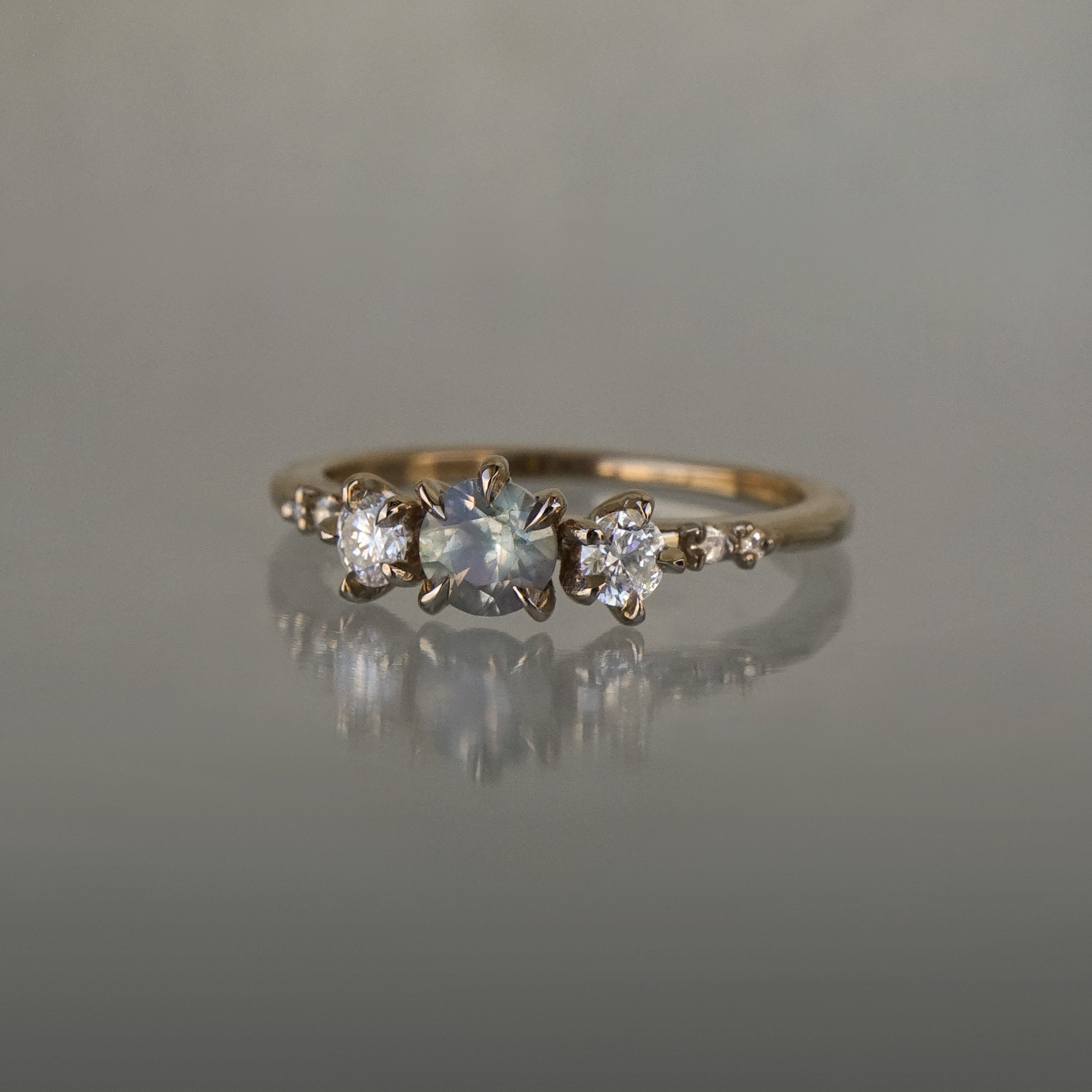 A one of a kind "Wild Iris" style engagement ring by Laurie Fleming Jewellery featuring a colour-change sapphire centre with shifting hues of blue, grey, blush, and green, flanked by a diamond on either side. Additional diamonds drip down the band. The ring is handcarved and made in solid champagne yellow gold, and is situated on a medium grey background.