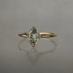A completely one of a kind, handcarved "Moondew" engagement ring by Laurie Fleming Jewellery. The ring features an oval Starbrite-cut colour change sapphire by lapidary artist John Dyer. The stone is held in four delicate claws, resting atop a gently tapered shining gold band. The ring is situated against a plain grey background.
