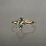 A completely one of a kind, handcarved "Moondew" engagement ring by Laurie Fleming Jewellery. The ring features an oval Starbrite-cut colour change sapphire by lapidary artist John Dyer. The stone is held in four delicate claws, resting atop a gently tapered shining gold band. The ring is situated against a plain grey background.