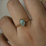 A completely one of a kind, handcarved "Moondew" engagement ring by Laurie Fleming Jewellery. The ring features an oval Starbrite-cut colour change sapphire by lapidary artist John Dyer. The stone is held in four delicate claws, resting atop a gently tapered shining gold band. The ring is worn on a hand against a plain light grey background.