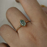 A completely one of a kind, handcarved "Moondew" engagement ring by Laurie Fleming Jewellery. The ring features an oval Starbrite-cut colour change sapphire by lapidary artist John Dyer. The stone is held in four delicate claws, resting atop a gently tapered shining gold band. The ring is worn on a hand against a plain light grey background.