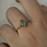 A completely one of a kind, handcarved "Moondew" engagement ring by Laurie Fleming Jewellery. The ring features an oval Starbrite-cut colour change sapphire by lapidary artist John Dyer. The stone is held in four delicate claws, resting atop a gently tapered shining gold band. The ring is worn on a hand against a plain light grey background.