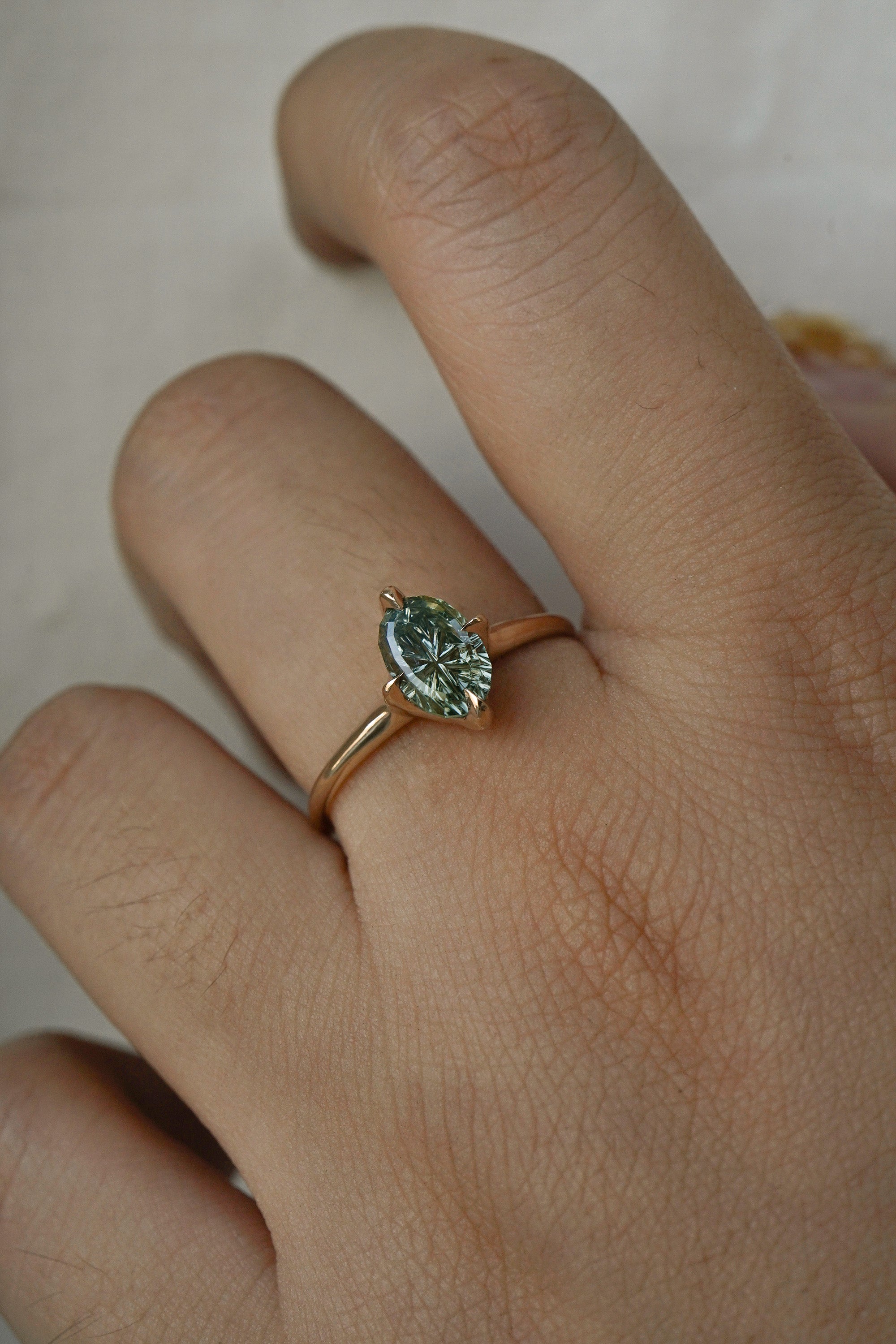 A completely one of a kind, handcarved "Moondew" engagement ring by Laurie Fleming Jewellery. The ring features an oval Starbrite-cut colour change sapphire by lapidary artist John Dyer. The stone is held in four delicate claws, resting atop a gently tapered shining gold band. The ring is worn on a hand against a plain light grey background.