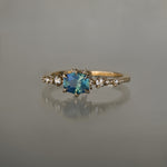 A delicate and ethereal "Nereid" one of a kind engagement ring by Laurie Fleming Jewellery, with a unique opalescent silky teal/turquoise/blue sapphire in a geometric oval cut. The band is adorned with rose and brilliant cut diamonds and the ring is made in solid 14k champagne gold. The ring is positioned against a medium grey background.