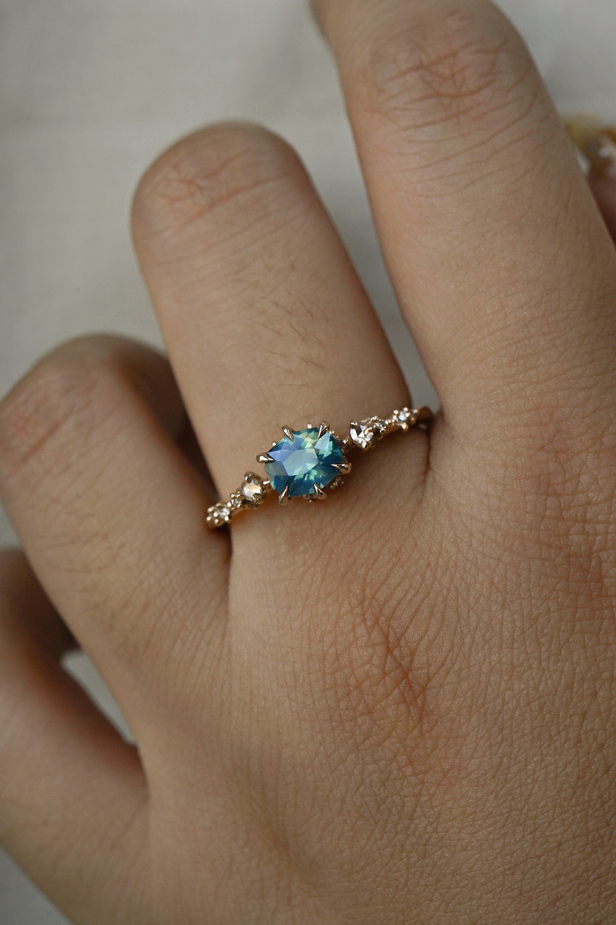 A delicate and ethereal "Nereid" one of a kind engagement ring by Laurie Fleming Jewellery, with a unique opalescent silky teal/turquoise/blue sapphire in a geometric oval cut. The band is adorned with rose and brilliant cut diamonds and the ring is made in solid 14k champagne gold. The ring is worn on a hand against a plain light grey background.