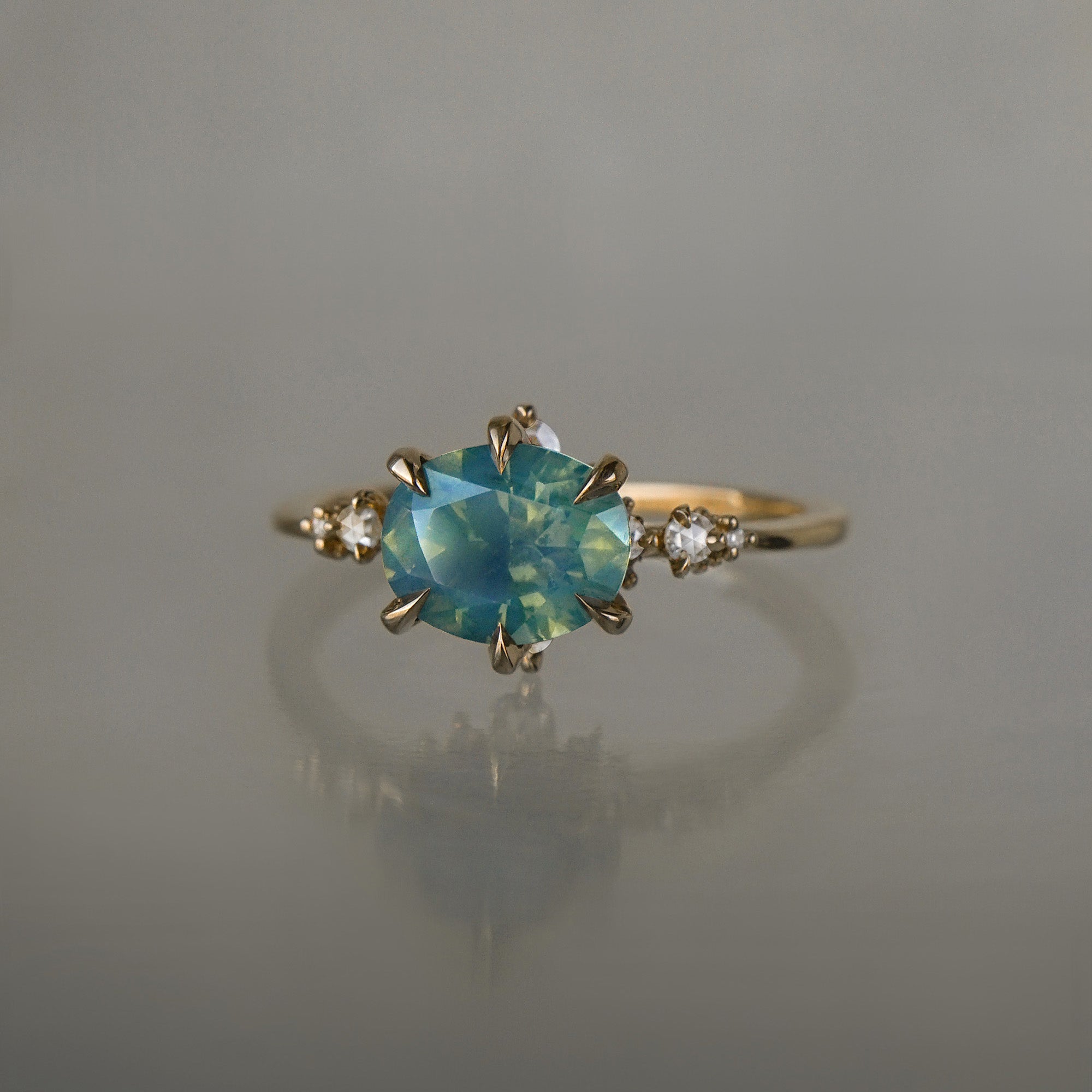 A delicate and ethereal "Nereid" one of a kind engagement ring by Laurie Fleming Jewellery, with a unique opalescent silky teal/turquoise/blue sapphire in an oval cut. The band is adorned with rose and brilliant cut diamonds and the ring is made in solid 14k yellow gold. The ring is positioned against a medium grey background.