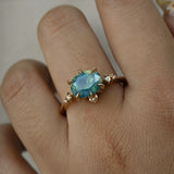 A delicate and ethereal "Nereid" one of a kind engagement ring by Laurie Fleming Jewellery, with a unique opalescent silky teal/turquoise/blue sapphire in an oval cut. The band is adorned with rose and brilliant cut diamonds and the ring is made in solid 14k yellow gold. The ring is worn on a hand against a plain lgiht grey background.