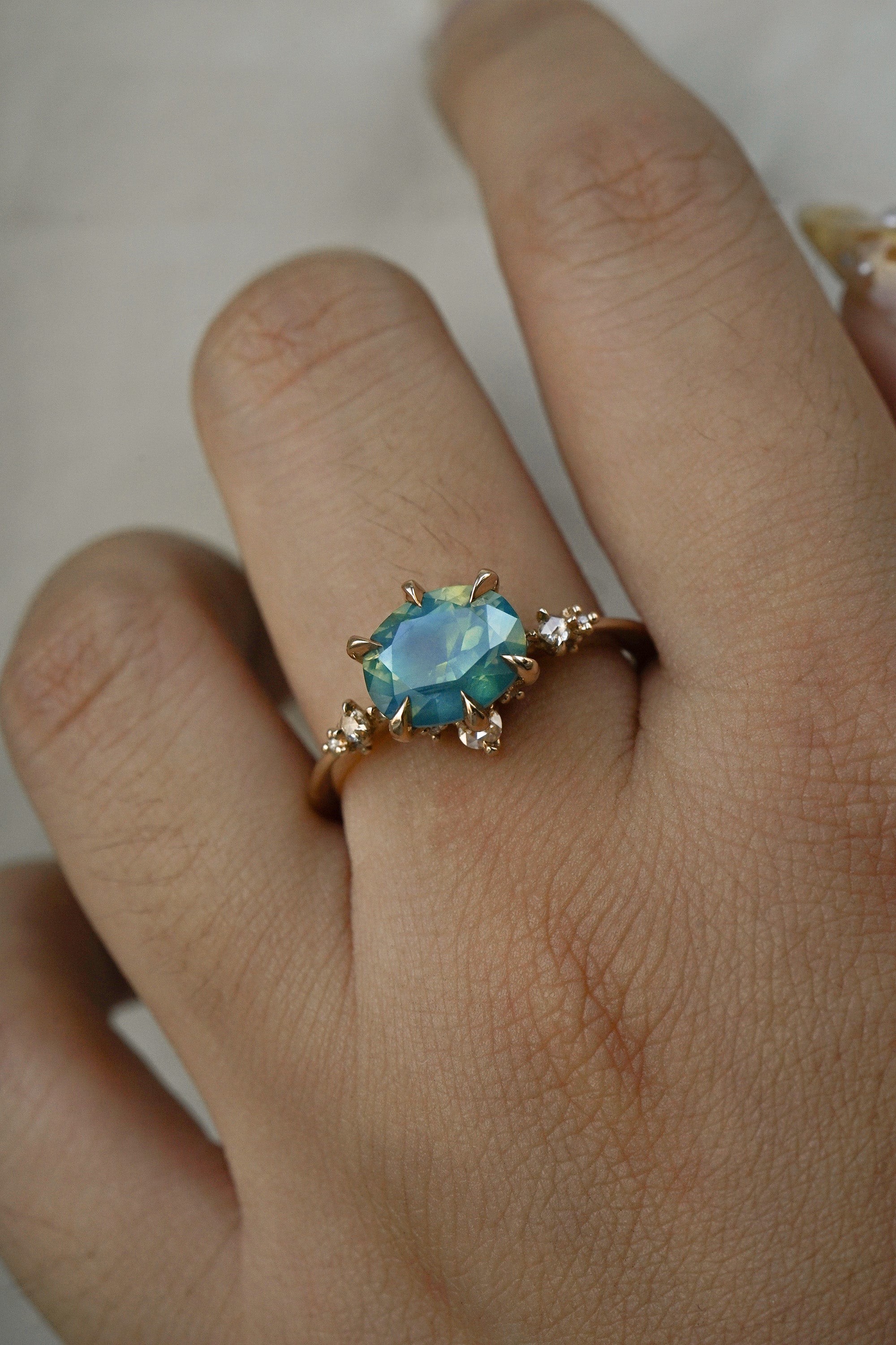 A delicate and ethereal "Nereid" one of a kind engagement ring by Laurie Fleming Jewellery, with a unique opalescent silky teal/turquoise/blue sapphire in an oval cut. The band is adorned with rose and brilliant cut diamonds and the ring is made in solid 14k yellow gold. The ring is worn on a hand against a plain lgiht grey background.