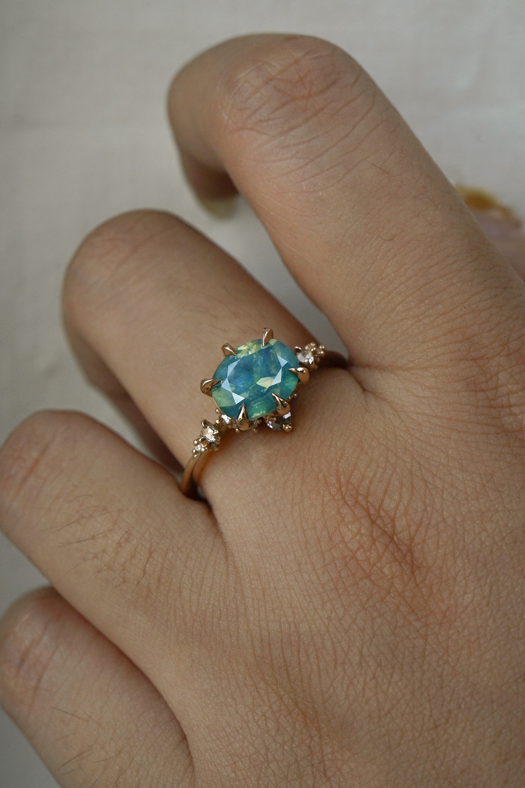 A delicate and ethereal "Nereid" one of a kind engagement ring by Laurie Fleming Jewellery, with a unique opalescent silky teal/turquoise/blue sapphire in an oval cut. The band is adorned with rose and brilliant cut diamonds and the ring is made in solid 14k yellow gold. The ring is worn on a hand against a plain light grey background.