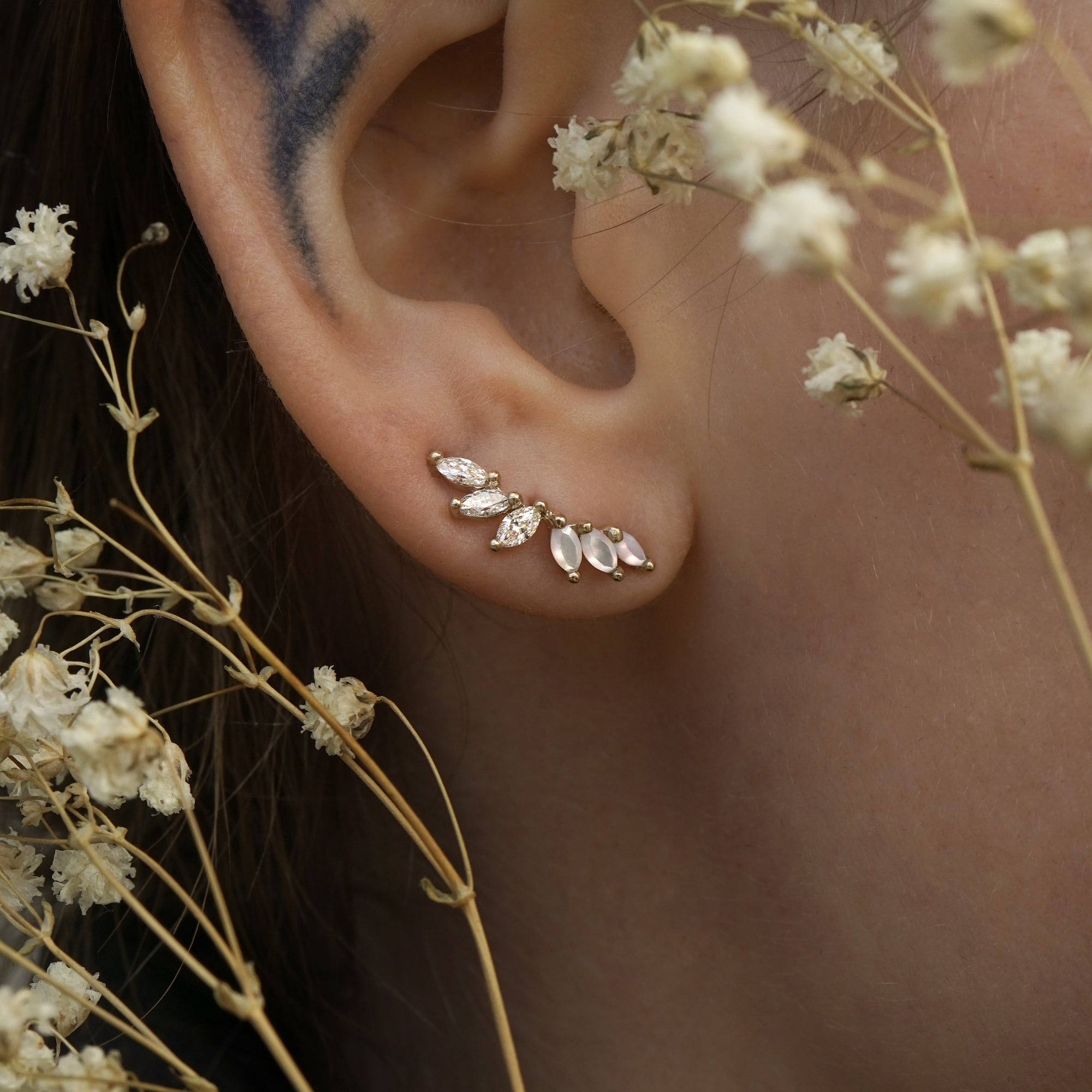 A close up of an ear wearing a Lyrie stud earring with three marquise cut mother of pearl and three marquise cut diamonds. There are dried flower stems in the foreground around the ear.