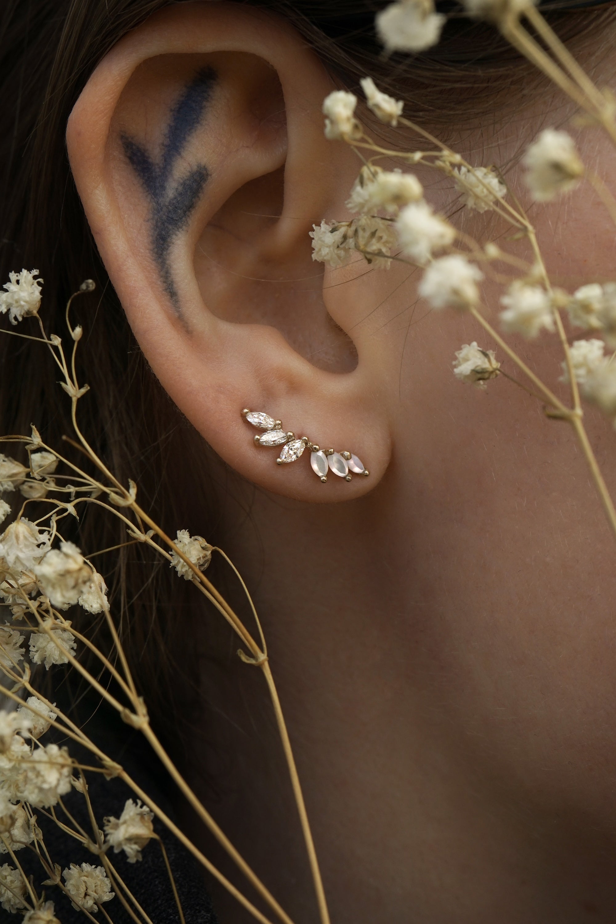 A close up of an ear wearing a Lyrie stud earring with three marquise cut mother of pearl and three marquise cut diamonds. There are dried flower stems in the foreground around the ear.