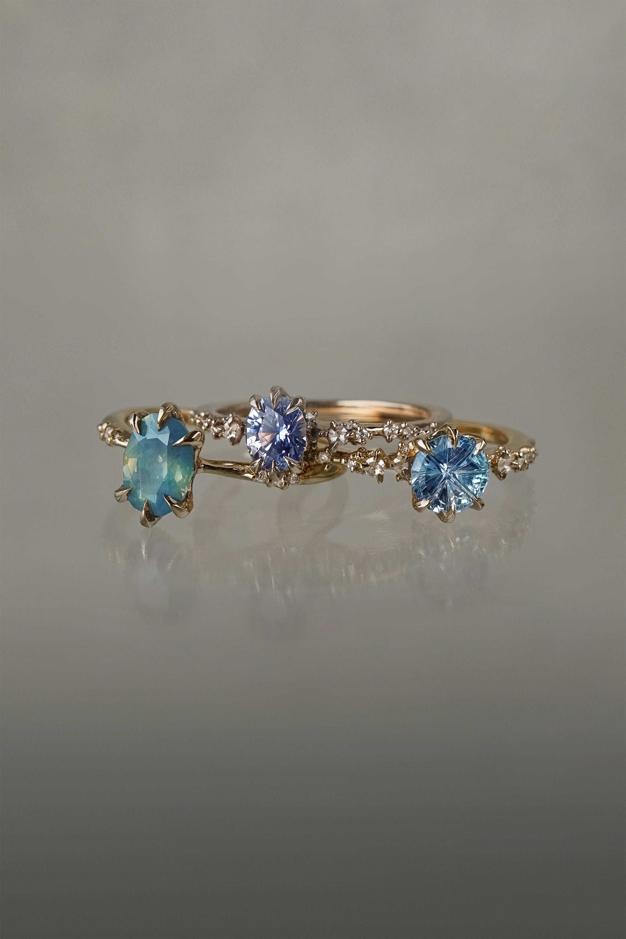 A group of three one of a kind rings by Laurie Fleming Jewellery. From left to right: An Ilona ring featuring an oval cut teal opalescent sapphire, a Nereid ring featuring an oval cut light blue purple pastel sapphire, and a Nereid ring with a John Dyer Starbrite cut blue sapphire centre. The rings are against a grey background.