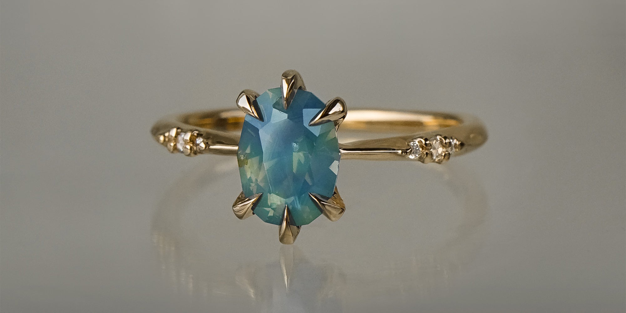 One of a kind "Ilona" ring by Laurie Fleming Jewellery featuring an oval cut opalescent turquoise teal sapphire with golden yellow sparkles. The ring has a delicate, tapered band with small diamond accents. It is in front of a medium grey background.