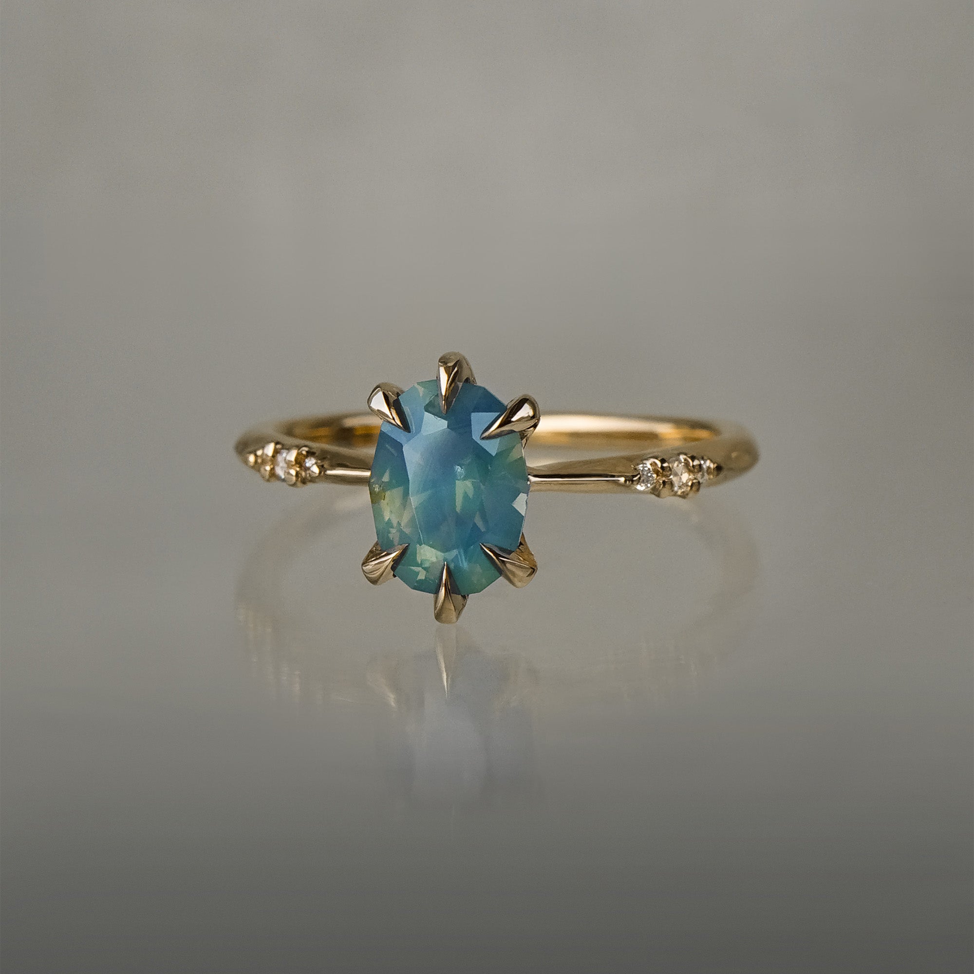 One of a kind "Ilona" ring by Laurie Fleming Jewellery featuring an oval cut opalescent turquoise teal sapphire with golden yellow sparkles. The ring has a delicate, tapered band with small diamond accents. It is in front of a medium grey background.