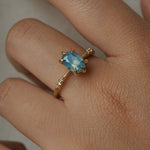 One of a kind "Ilona" ring by Laurie Fleming Jewellery featuring an oval cut opalescent turquoise teal sapphire with golden yellow sparkles. The ring has a delicate, tapered band with small diamond accents. The ring is worn on a hand.