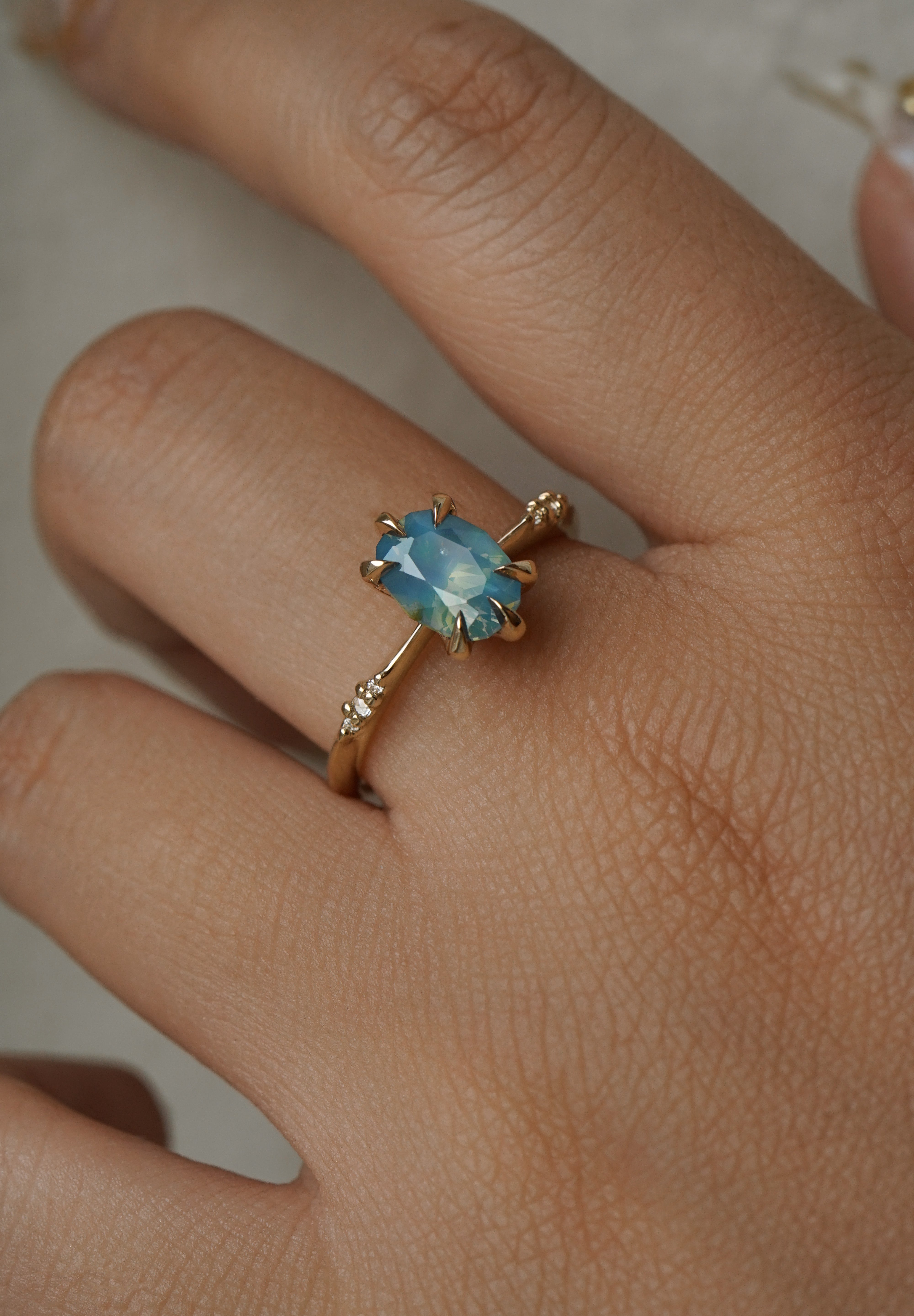 One of a kind "Ilona" ring by Laurie Fleming Jewellery featuring an oval cut opalescent turquoise teal sapphire with golden yellow sparkles. The ring has a delicate, tapered band with small diamond accents. The ring is worn on a hand.