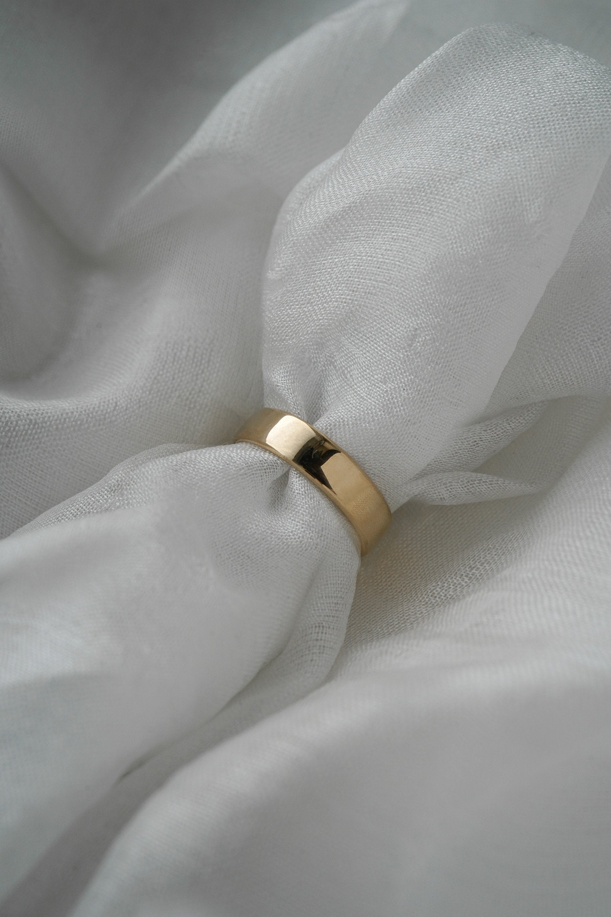 A stock photo of Laurie Fleming Jewellery's plain solid gold "Juniper" band, a 5mm wide flat comfort fit stacking ring/wedding band, handmade in Toronto. The ring is held on a roll of white silk fabric which is nestled against more silk.