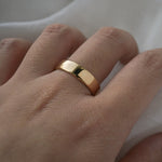 A stock photo of Laurie Fleming Jewellery's plain solid gold "Juniper" band, a 5mm wide flat comfort fit stacking ring/wedding band, handmade in Toronto. The ring is worn on a hand with a light grey background.