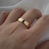 A stock photo of Laurie Fleming Jewellery's plain solid gold "Juniper" band, a 5mm wide flat comfort fit stacking ring/wedding band, handmade in Toronto. The ring is worn on a hand with a light grey background.