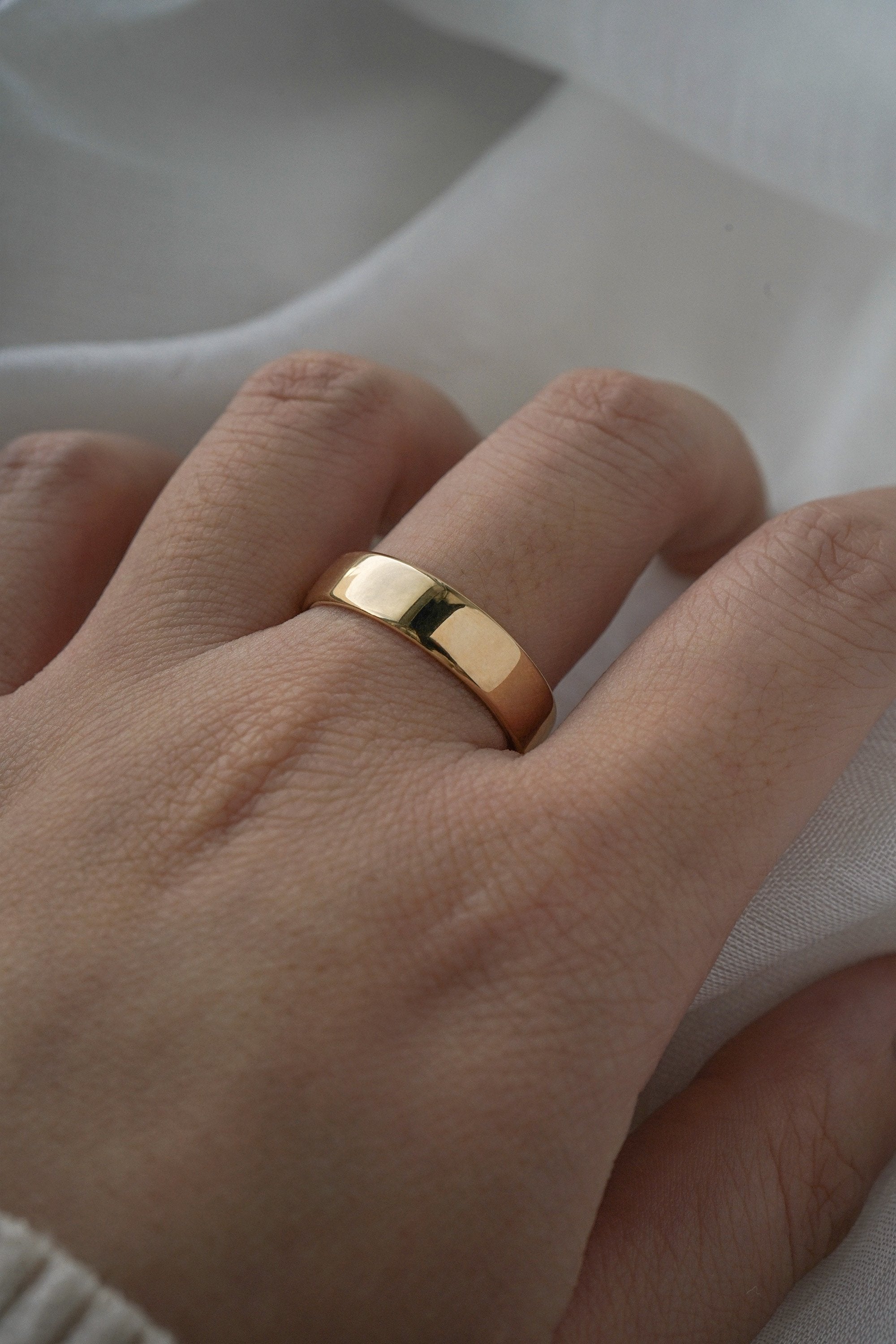 A stock photo of Laurie Fleming Jewellery's plain solid gold "Juniper" band, a 5mm wide flat comfort fit stacking ring/wedding band, handmade in Toronto. The ring is worn on a hand with a light grey background.