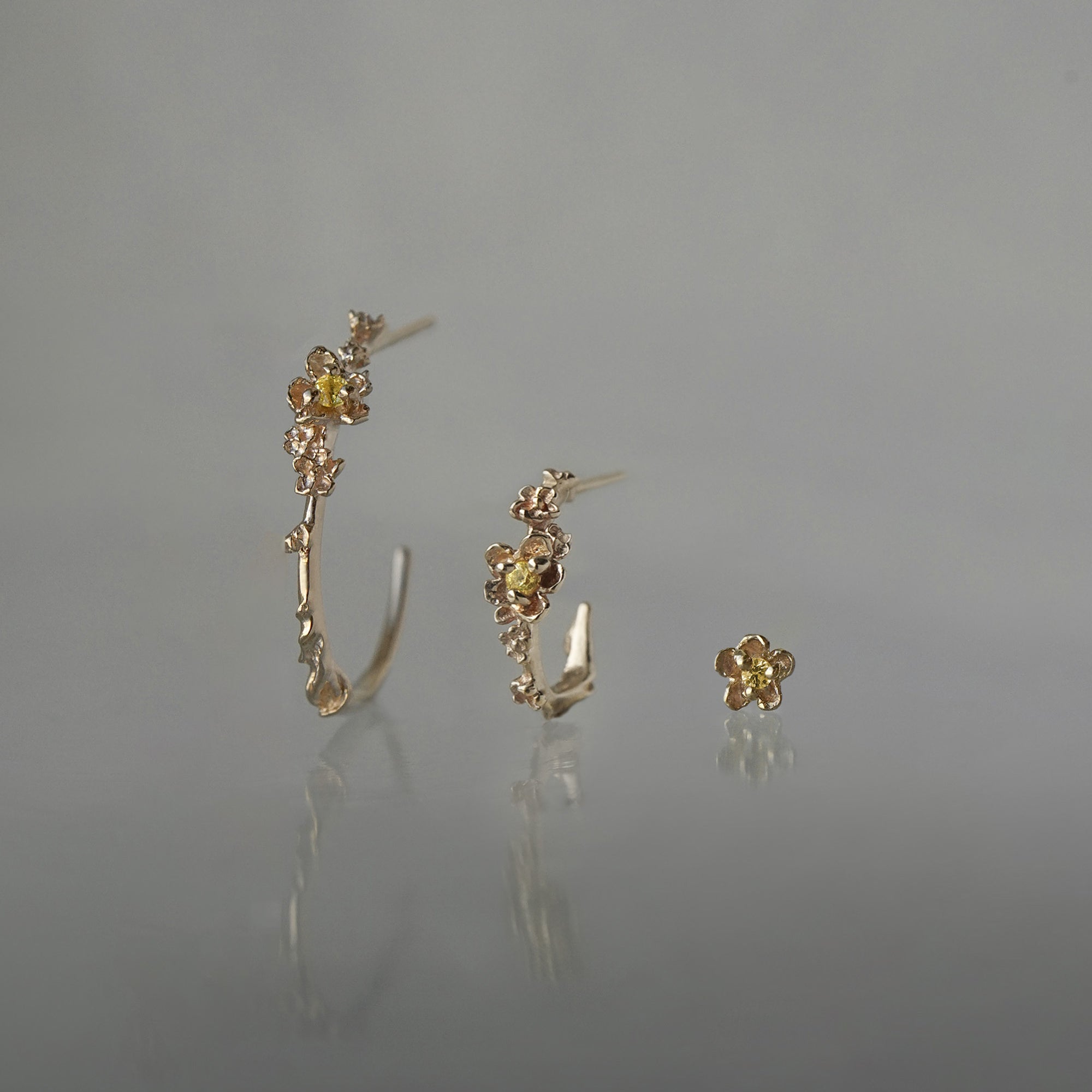 A line-up of three Laurie Fleming Jewellery earrings over a medium grey background. On the left, a large buttercup hoop. In the middle,  a small buttercup hoop. On the right, a buttercup stud earring. All three earrings feature hand-carved petite buttercup flower blossoms with yellow sapphire centres.