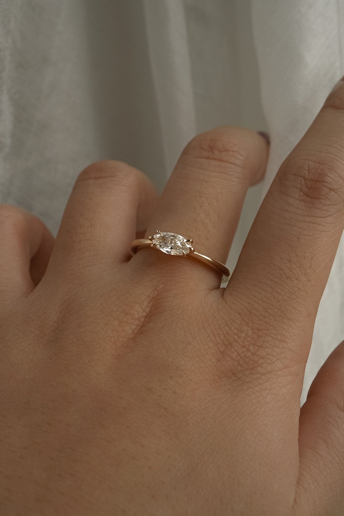 A one of a kind "Aurelia" style engagement ring by Laurie Fleming Jewellery, featuring a unique "moval" (marquise/oval hybrid) cut diamond set East-West, on a gently tapered and rounded solid gold band. The ring is worn on a hand that is posed in front of some draped gossamer silk fabric.