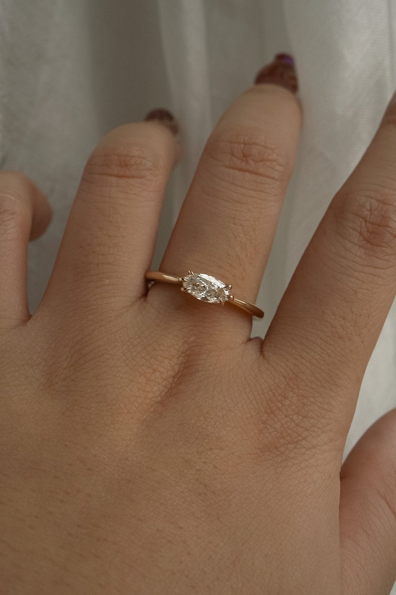 A one of a kind "Aurelia" style engagement ring by Laurie Fleming Jewellery, featuring a unique "moval" (marquise/oval hybrid) cut diamond set East-West, on a gently tapered and rounded solid gold band. The ring is worn on a hand that is posed in front of some draped gossamer silk fabric.