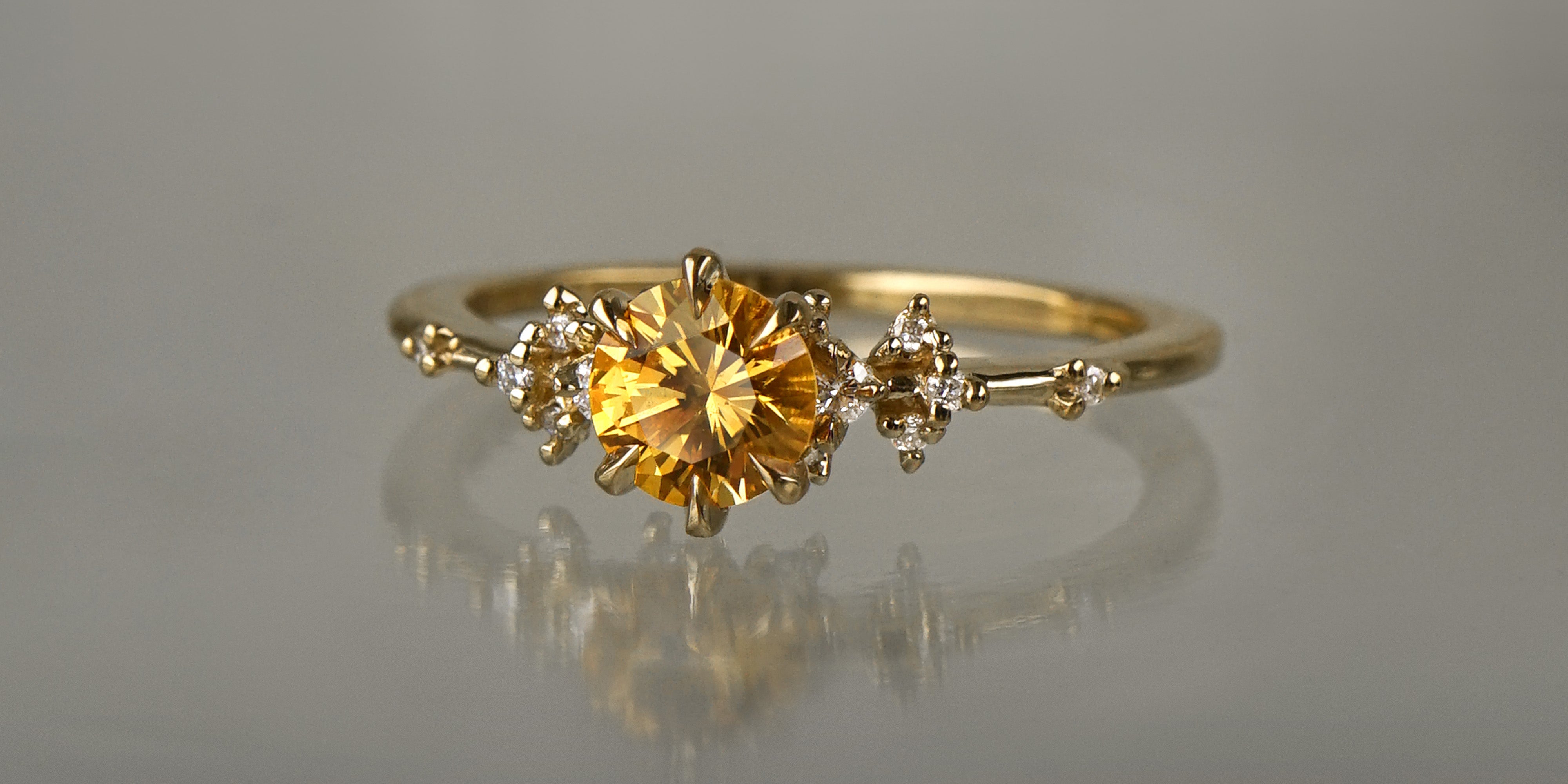 A one of a kind 18k yellow gold Water Lily engagement ring by Laurie Fleming Jewellery with a bright golden yellow round Montana sapphire centre with clusters of diamonds on the band. The ring is situated on a medium grey background.